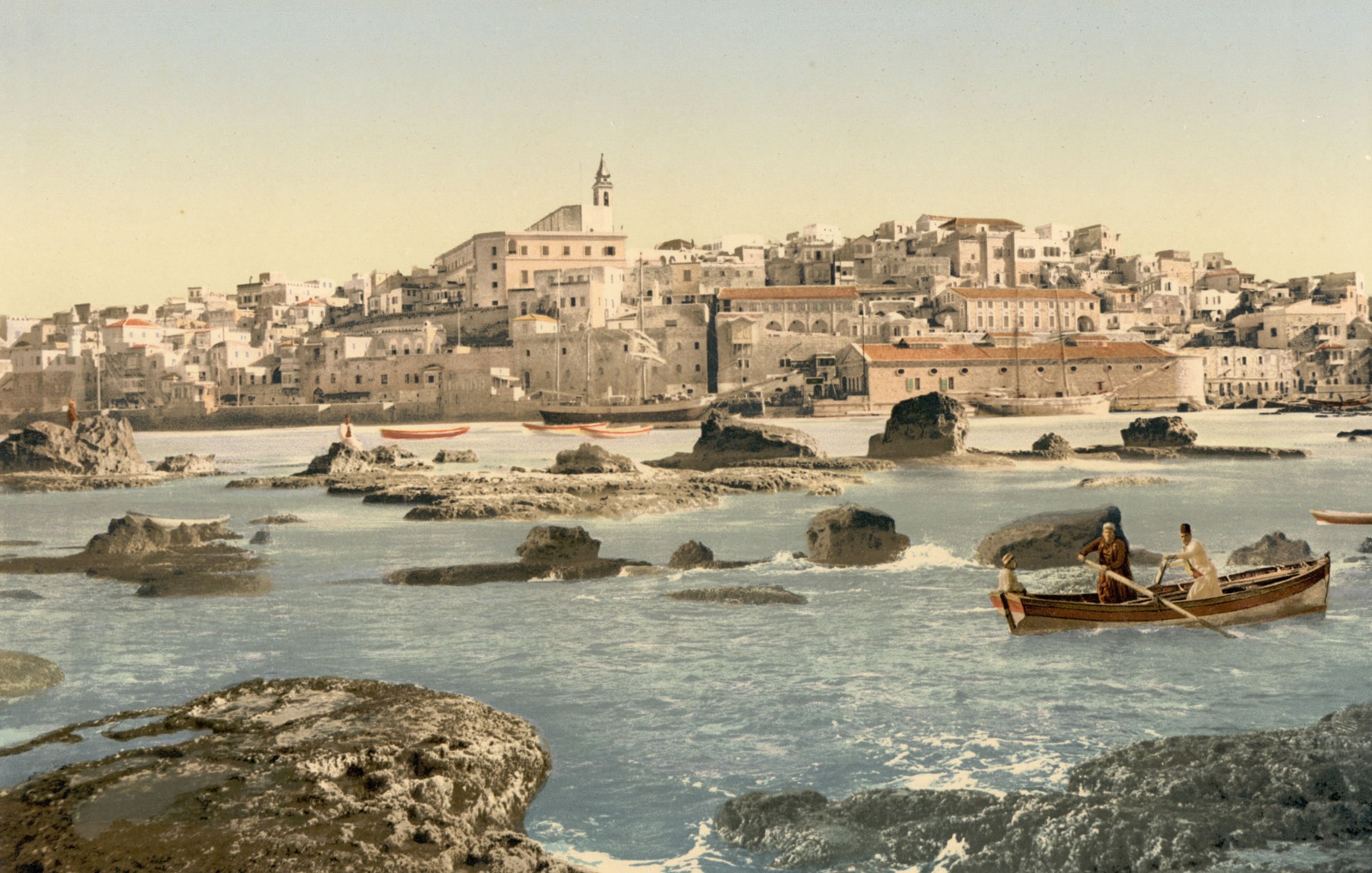 L'image représente un paysage côtier avec une ville en arrière-plan. On peut voir des maisons blanches construisant un front de mer, accompagnées d'une église au sommet d'une colline. Au premier plan, deux personnes naviguent dans un petit bateau sur une eau calme, entouré de rochers. Le ciel est dégagé, suggérant une journée ensoleillée, et la scène évoque une atmosphère paisible et pittoresque. Les couleurs sont vives, mettant en valeur la beauté naturelle de l'endroit.