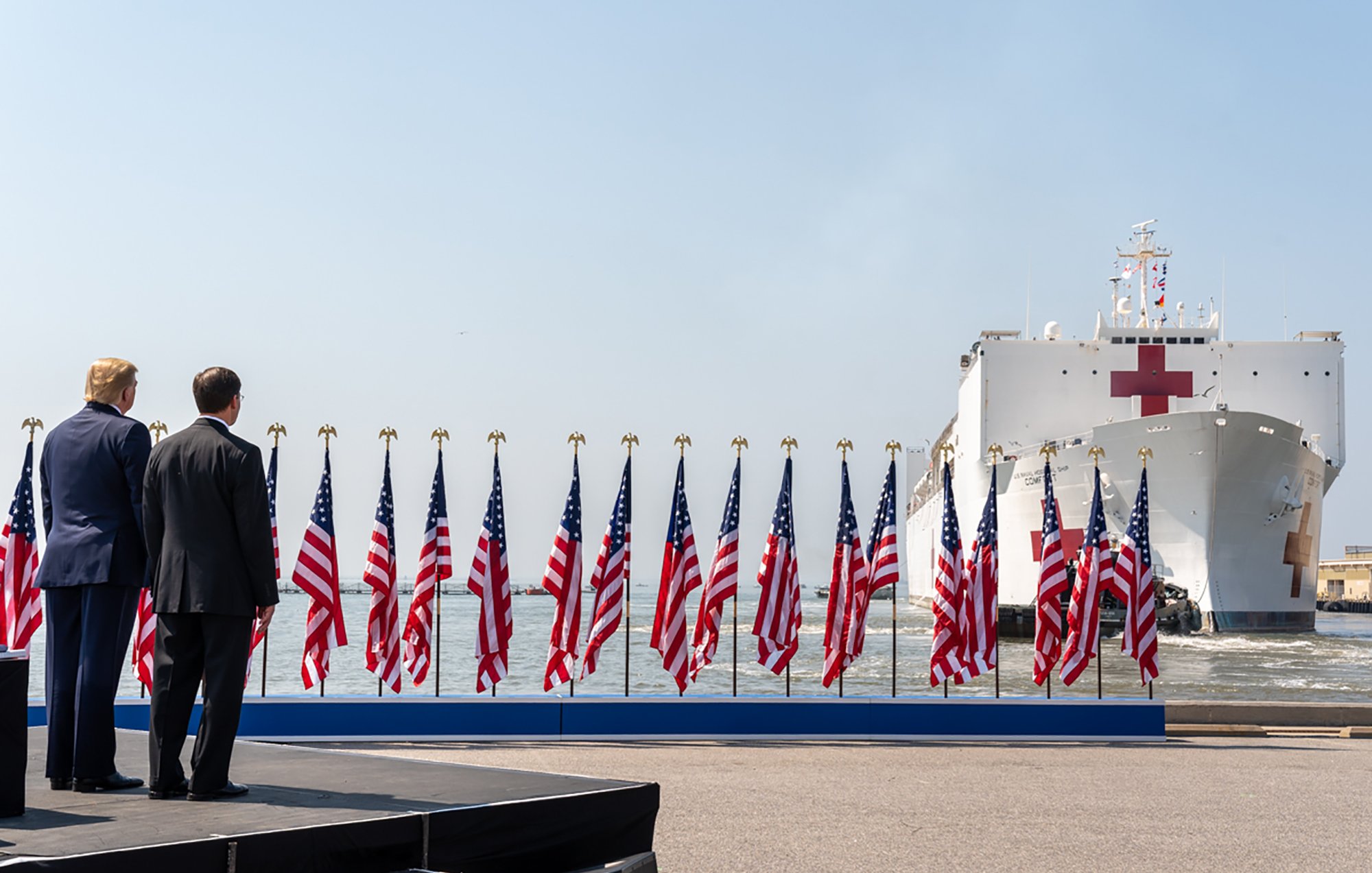 L'image montre une scène avec deux hommes de dos, face à un grand navire hospitalier qui arrive. Le navire est identifiable grâce à sa croix rouge sur le côté. Au premier plan, un podium est visible, entouré de multiples drapeaux américains flottant au vent. Le ciel est clair et lumineux, suggérant une journée ensoleillée. L'atmosphère semble solennelle, probablement en lien avec un événement officiel ou une cérémonie.