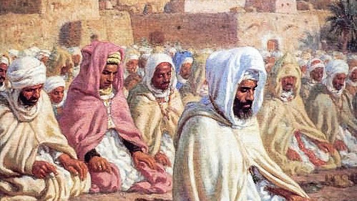 L'image représente un groupe de personnes rassemblées en prière, probablement dans un contexte religieux. Elles sont vêtues de robes traditionnelles et portent des turbans ou des voiles. Les hommes semblent concentrés et méditatifs, assis sur le sol. En arrière-plan, on peut voir une ville avec des maisons aux couleurs claires, sous un ciel bleu, ce qui suggère un environnement désertique ou méditerranéen. L'atmosphère est paisible et spirituelle.