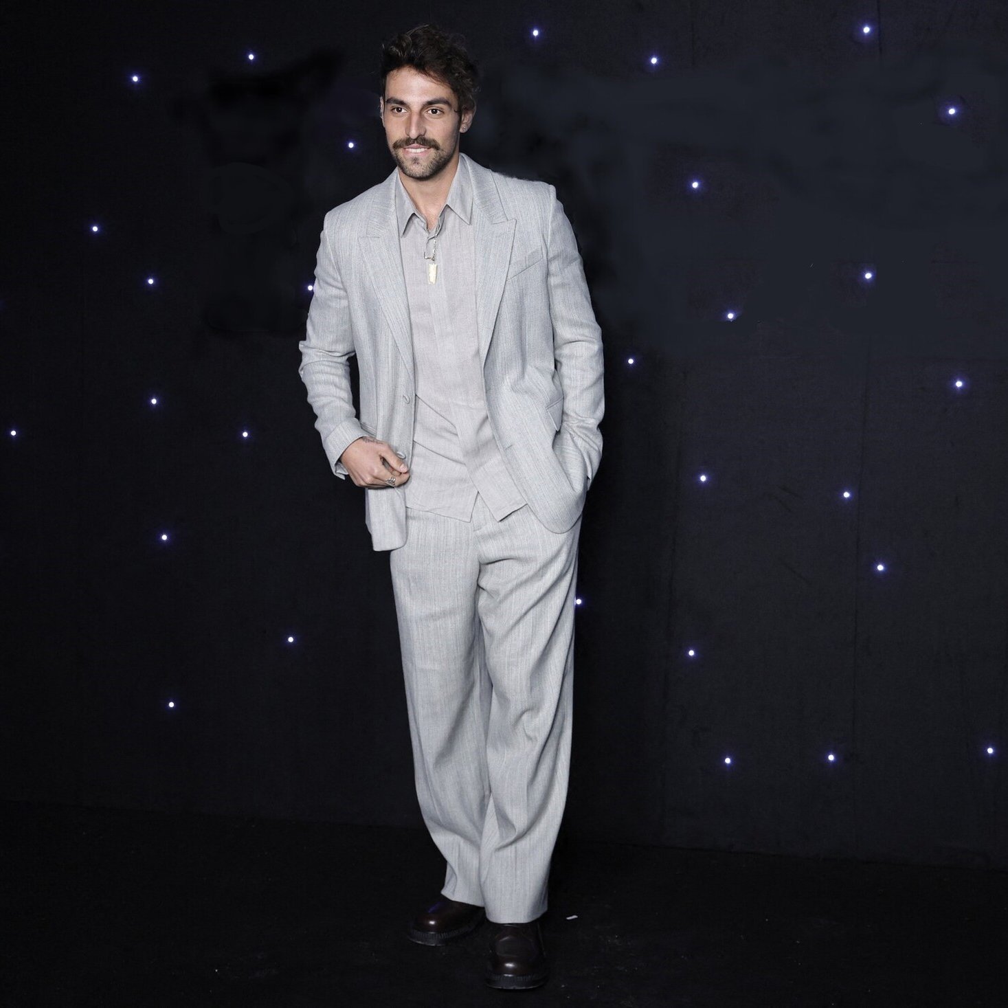 L'image montre un homme portant un costume léger à rayures, composé d'une veste et d'un pantalon ample. Il a une moustache et un sourire, et se tient debout avec une pose décontractée. L'arrière-plan est sombre, parsemé de petites lumières blanches qui ajoutent un effet élégant à la scène.