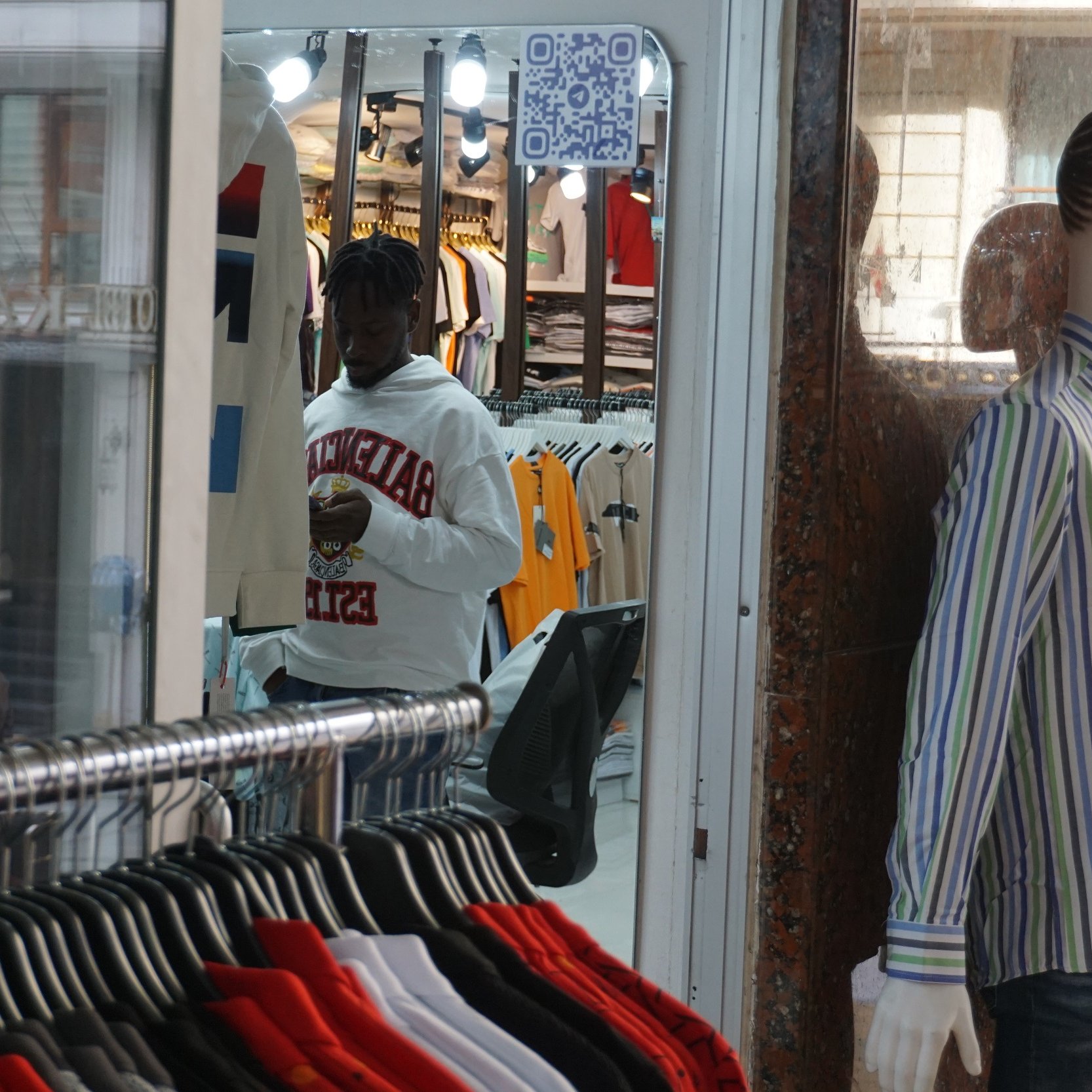 L'image montre un magasin de vêtements avec des rangées de vêtements accrochés, notamment des t-shirts et des chemises multicolores. À gauche, un homme portant un sweat-shirt blanc est en train de regarder son téléphone. À droite, un mannequin habillé d'une chemise à rayures est exposé dans la vitrine. On peut aussi apercevoir un code QR accroché à l'intérieur du magasin. L ambiance est typique d'un espace commercial animé.
