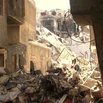 L'image montre une scène de destruction, probablement dans une zone urbaine touchée par un conflit ou un désastre. Des bâtiments sont en ruines, avec des murs effondrés et des débris éparpillés sur le sol. On peut voir des tas de gravats et de déchets, ce qui suggère que la zone a été abandonnée ou négligée. La lumière du soleil pénètre à travers les décombres, créant un contraste entre l'ombre et la lumière. L'atmosphère évoque la désolation et la tristesse.