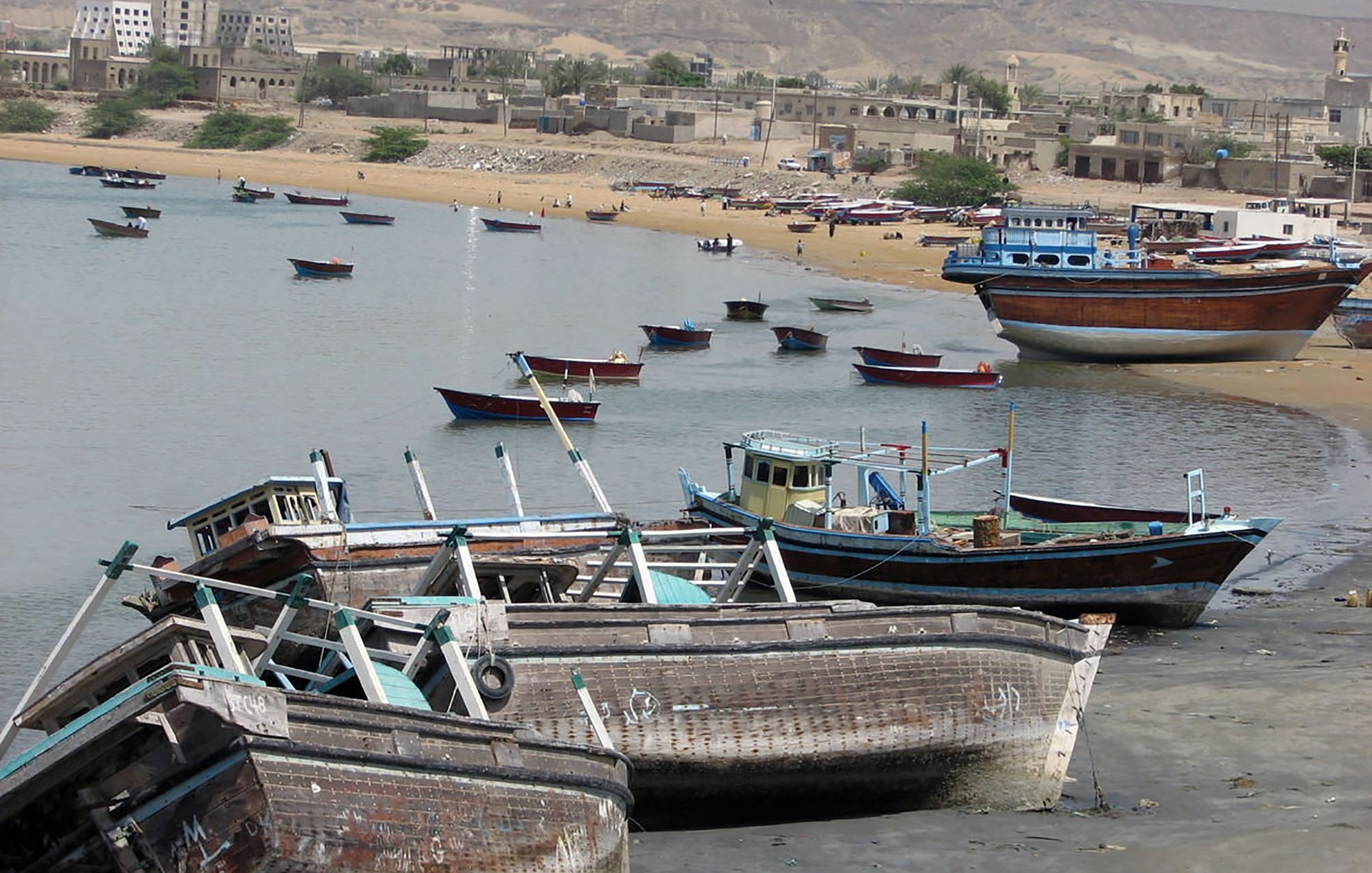 L'image montre une scène côtière avec plusieurs bateaux de pêche échoués sur la plage. On peut voir des petites embarcations peintes en différentes couleurs, certaines abîmées, qui sont disposées le long d'une côte sablonneuse. En arrière-plan, des bâtiments témoignent d'une ville ou d'un village, et le paysage est dominé par des collines ou des falaises. L'eau est calme, et l'atmosphère semble tranquille, avec quelques personnes visibles sur la plage.