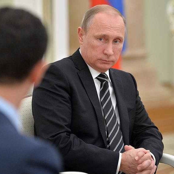 در تصویر، ولادیمیر پوتین در یک جلسه رسمی نشسته است. او لباسی تیره و رسمی بر تن دارد و به نظر می‌رسد در حال تفکر یا گوش دادن به سخنانی است که توسط فردی دیگر بیان می‌شود. زاویه‌ای از چهره‌ی فرد دیگری هم در سمت چپ تصویر دیده می‌شود، اما مشخصات او قابل شناسایی نیست. پس‌زمینه تصویر به سبک رسمی و متناسب با یک جلسه سیاسی طراحی شده است.