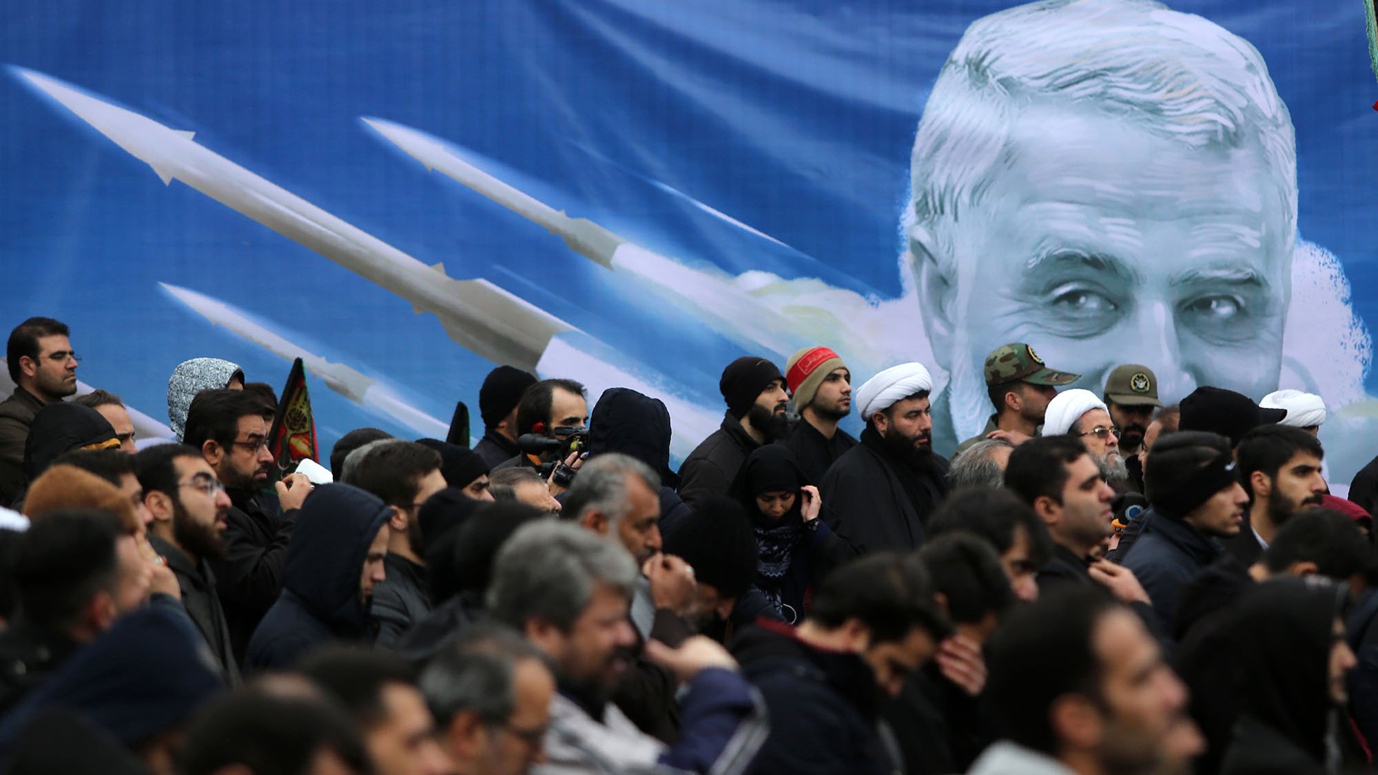 تصویر نشان‌دهنده تجمعی از افراد در یک مراسم است. مردم بیشتر لباس‌های تیره پوشیده‌اند و برخی از آن‌ها در حال گوش دادن به سخنان یا تماشای یک برنامه هستند. در پس‌زمینه، تصویری بزرگ از چهره‌ای که شبیه یک شخصیت شناخته‌شده است وجود دارد و در اطراف آن تصاویری از موشک‌ها دیده می‌شود. این تصویر به نوعی نمادین به نظر می‌رسد و می‌تواند به مناسبت‌های ملی یا مذهبی مربوط باشد. 