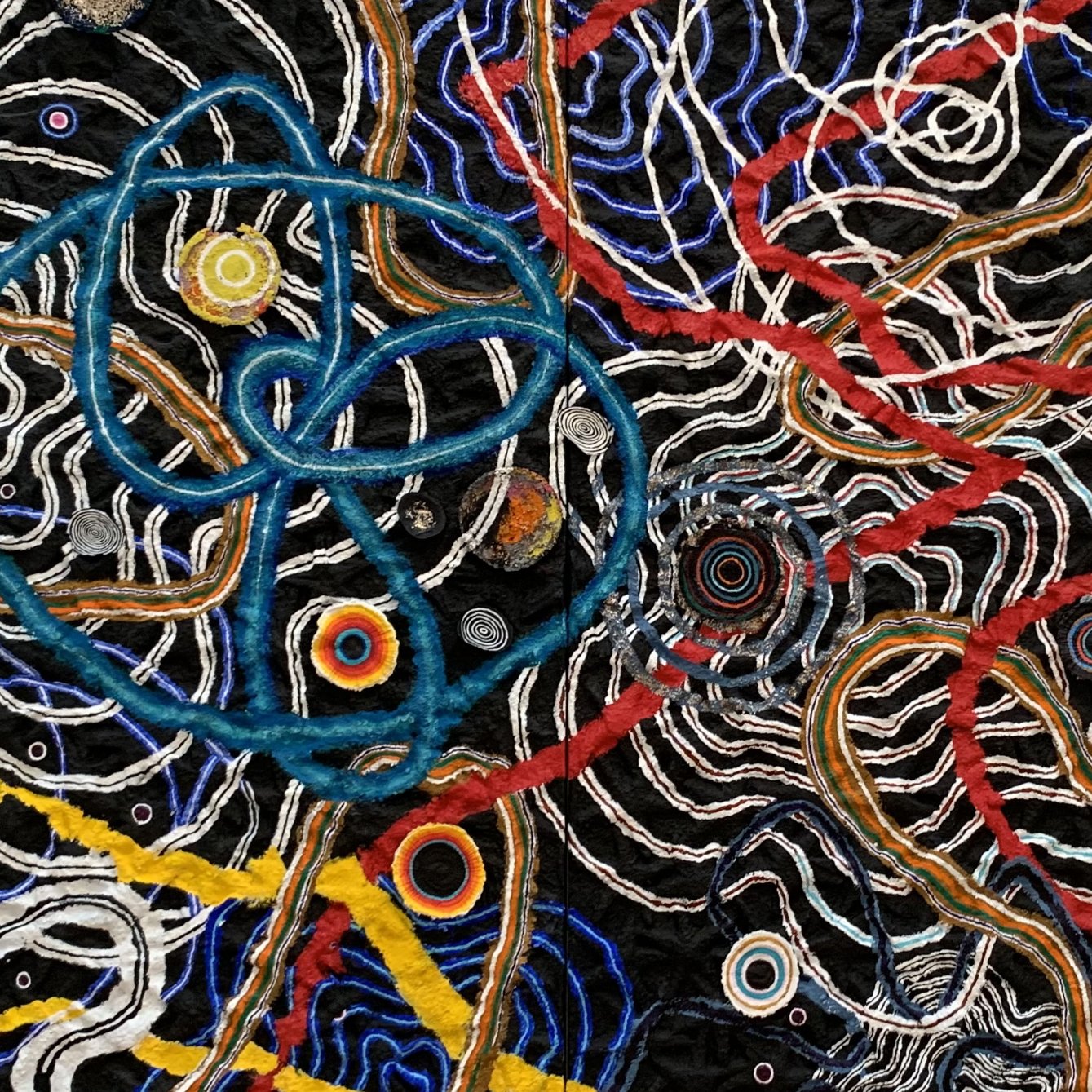 The image features a vibrant abstract design with intertwining lines and shapes. It includes a variety of colors, such as blue, red, yellow, and white, creating a dynamic and complex pattern. The background is dark, which enhances the brightness of the interwoven elements. Circular forms are scattered throughout, contributing to the overall sense of movement and depth, resembling a blend of organic and geometric motifs. The texture suggests a tactile quality, inviting viewers to explore the intricate connections within the composition.