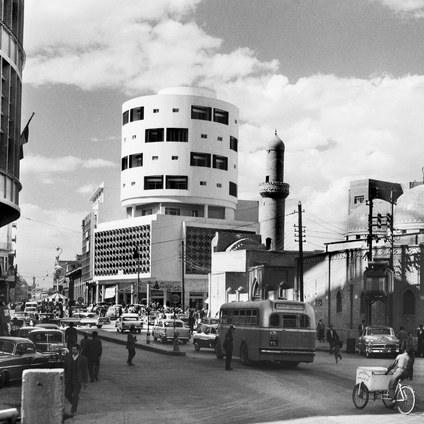 L'image en noir et blanc présente une scène urbaine animée. Au premier plan, on peut voir une rue avec plusieurs véhicules, dont des voitures et un bus. Le bâtiment à gauche a une architecture moderne, tandis qu'à droite, on aperçoit un édifice avec des dômes caractéristiques, suggérant une influence architecturale traditionnelle. Des piétons circulent, et on remarque également un homme sur un vélo. Le ciel est partiellement nuageux, ajoutant à l'atmosphère de la scène. Cette image évoque un mélange d'architecture moderne et traditionnelle dans un contexte urbain.