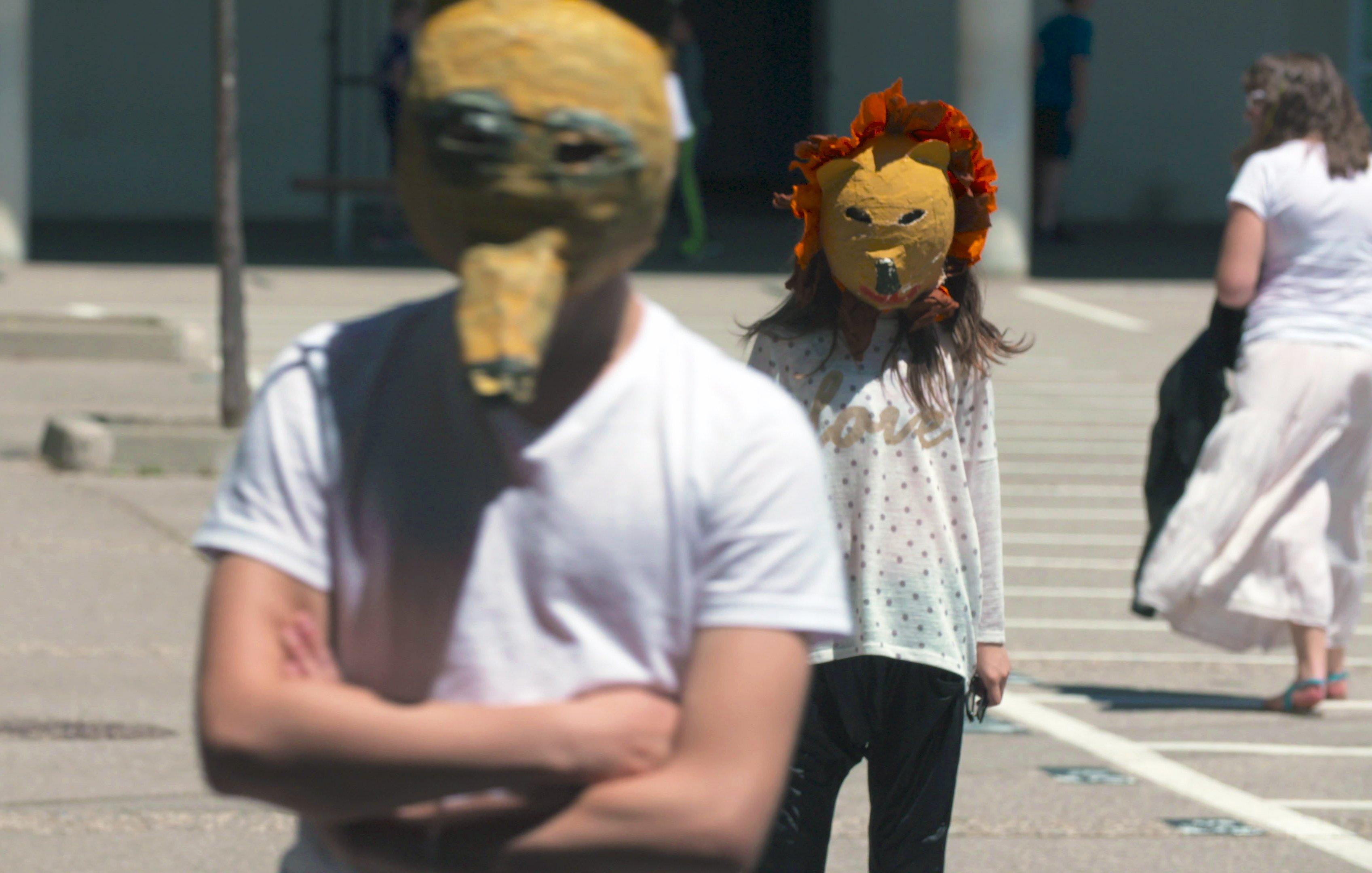 L'image montre deux personnes portant des masques originaux. L'un des masques a une forme de tête d'animal, tandis que l'autre est décoré de fleurs, évoquant un lion. Les personnes semblent se promener dans un espace extérieur, peut-être une place ou une cour. L'environnement est lumineux et rappelle une scène de performance artistique ou d'un événement communautaire.