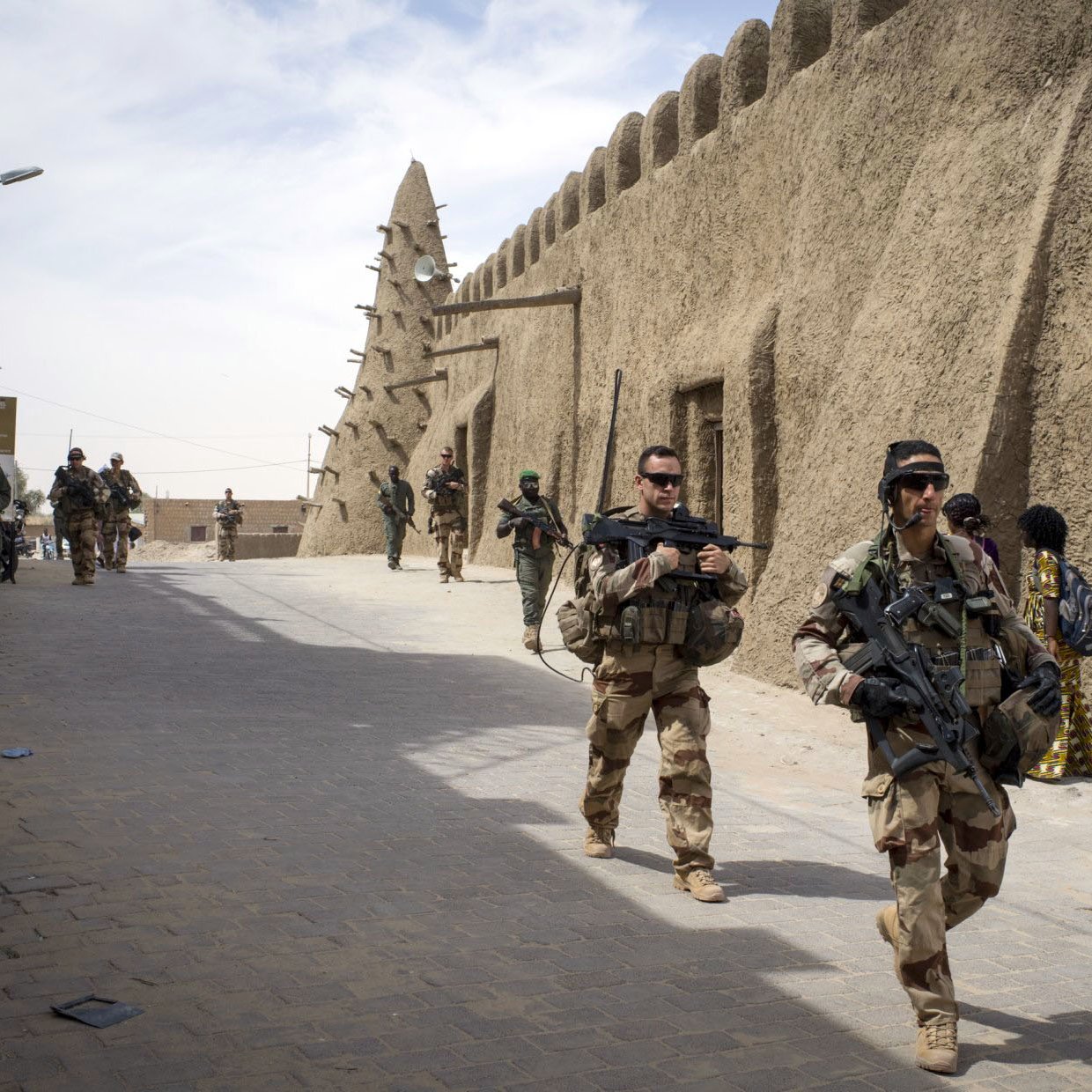 تظهر الصورة جنوداً يسيرون في شوارع منطقة تاريخية. المباني في الخلفية تبدو مصنوعة من الطين، وتمتاز بتصميم تقليدي. الجنود يحملون أسلحة ويبدو عليهم التركيز والحذر. يتواجد أيضاً شخصيات أخرى في الخلفية، مما يدل على وجود نشاطات متعددة في المنطقة. السماء تبدو صافية مع بعض السحب.