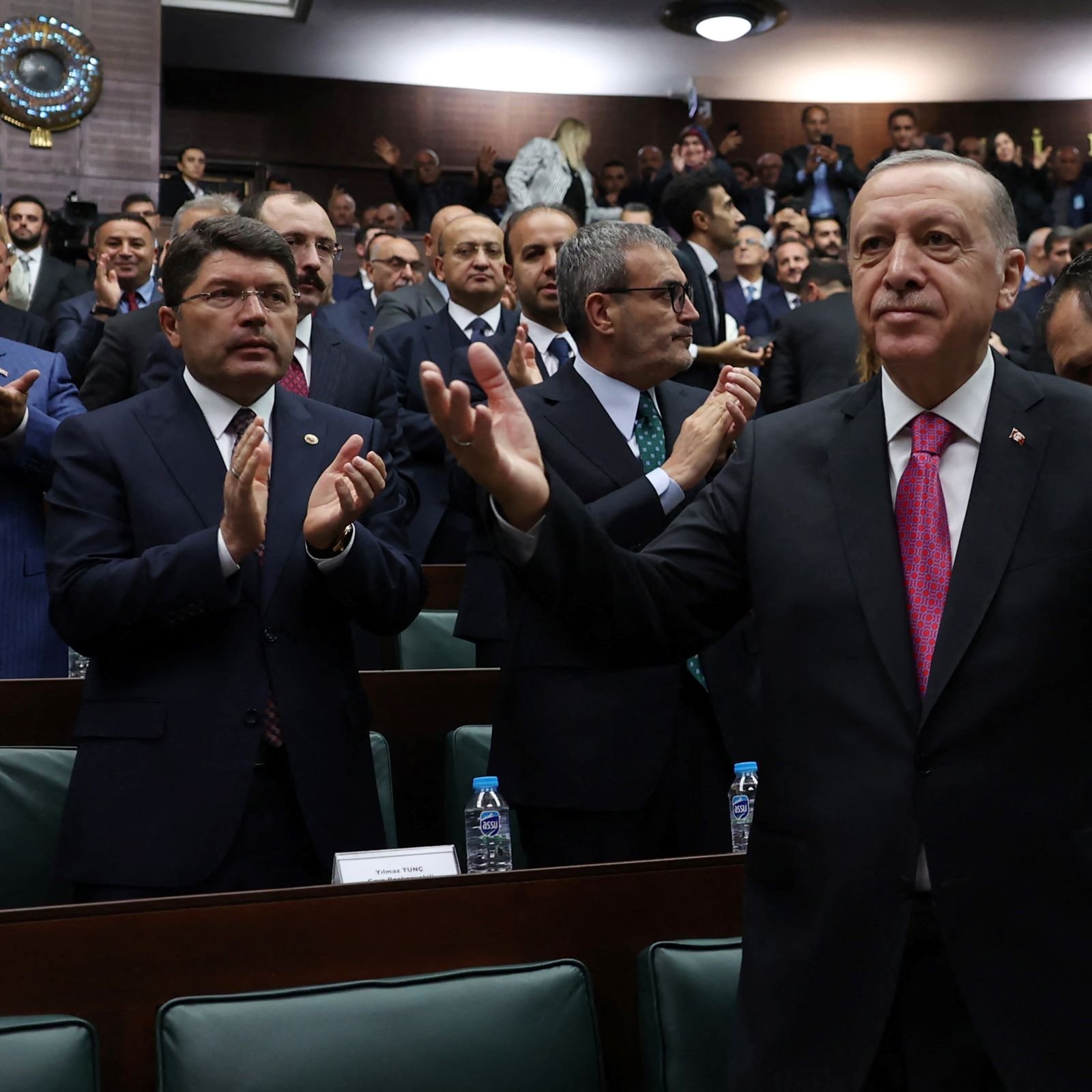 در این تصویر، یک جمعیت بزرگ از افرادی که در یک سالن جلسه نشسته‌اند، دیده می‌شود. در مرکز تصویر، فردی با لباس رسمی و کراوات صورتی در حال خوش‌آمدگویی یا ابراز احساسات است. او با دستان باز به سمت مخاطبان اشاره می‌کند. در پس‌زمینه، اعضای دیگر نیز ایستاده و به او نگاه می‌کنند. برخی از آنها در حال تشویق و applauding هستند. فضای کلی مراسم جدی و رسمی به نظر می‌رسد.