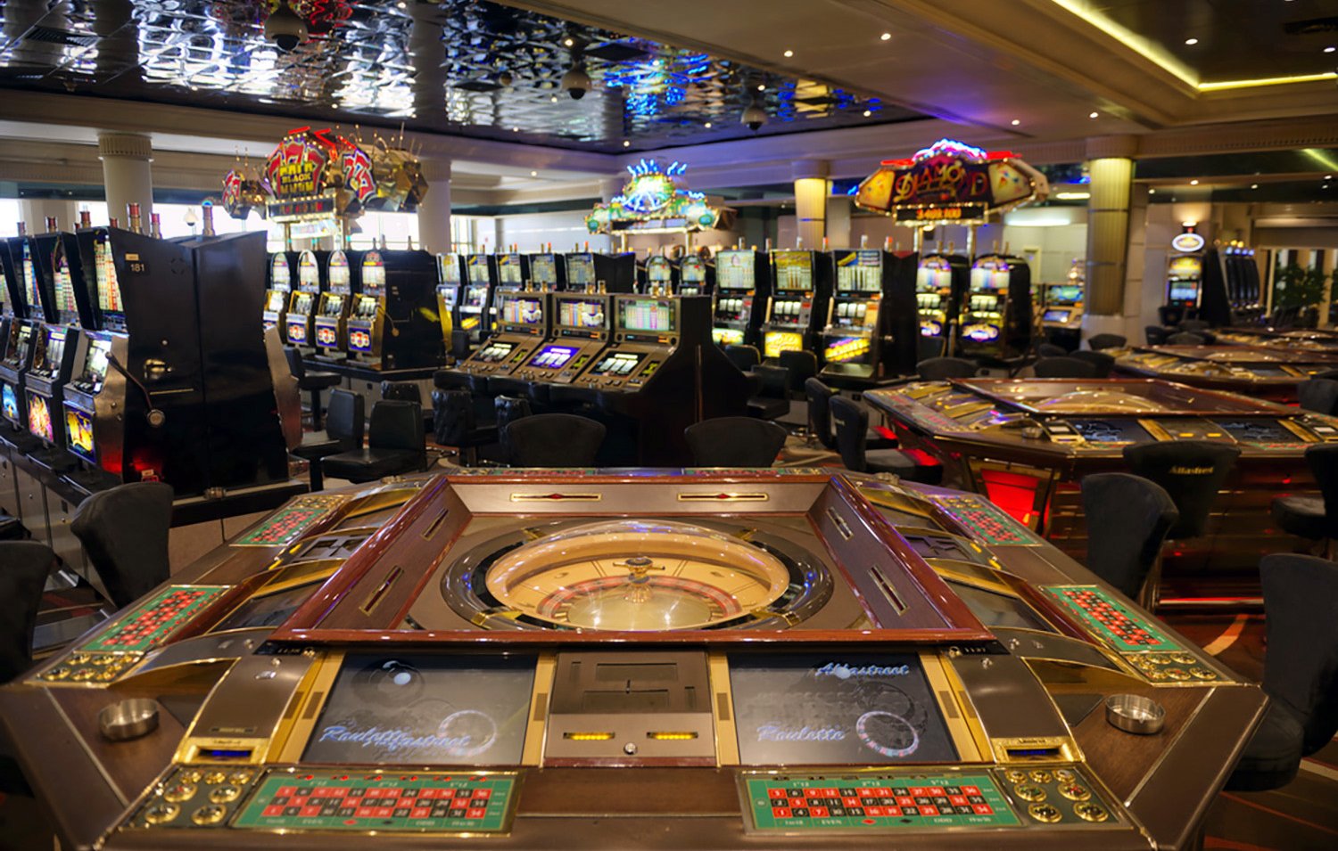 L'image montre l'intérieur d'un casino, avec plusieurs tables de jeux et des machines à sous. Au premier plan, on peut voir une grande table de roulette, entourée de plusieurs sièges. À l'arrière-plan, on distingue de nombreuses machines à sous illuminées, créant une atmosphère animée et colorée. Le plafond est décoré, ajoutant à l'élégance de l'espace. L'ensemble dégage une ambiance de divertissement et de luxe.