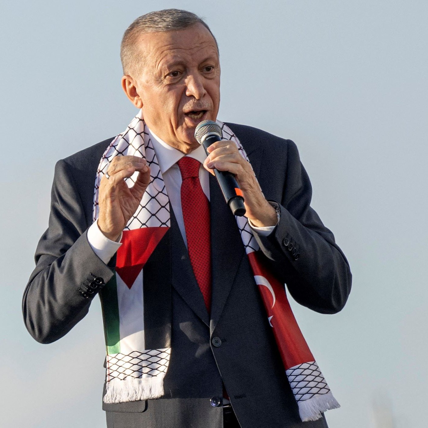 L'image montre un homme en train de parler au micro, probablement lors d'un discours ou d'une manifestation publique. Il porte un costume sombre avec une cravate rouge et un foulard aux motifs représentant la Palestine, qui est enroulé autour de son cou. L'arrière-plan semble clair, suggérant qu'il se trouve à l'extérieur. L'homme semble exprimer des émotions fortes, ce qui indique qu'il est engagé dans son discours.