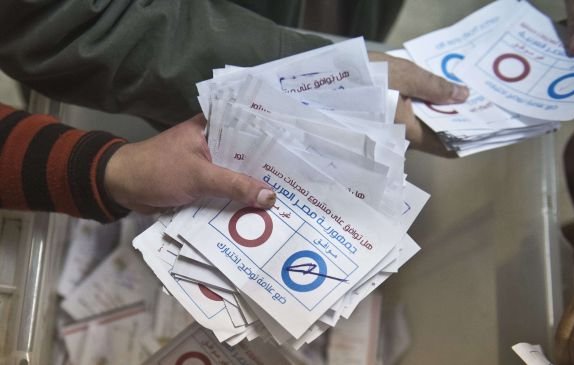 L'image montre des mains tenant des bulletins de vote. On peut voir des bulletins blancs portant des cercles rouges et bleus, ainsi que des inscriptions en arabe. Il semble s'agir d'une scène de vote, probablement lors d'une élection. Les bulletins sont rassemblés, suggérant un processus électoral en cours.