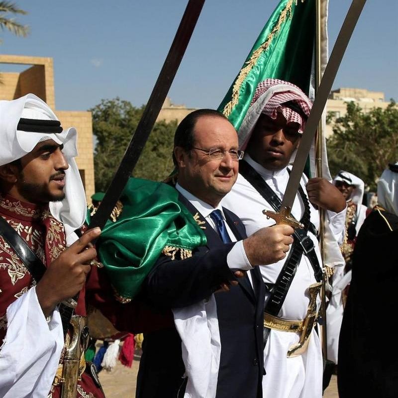 L'image montre un homme au centre, portant une veste sombre, accompagné de deux personnes de chaque côté. Ces dernières sont vêtues d'habits traditionnels arabes et tiennent des épées. L'arrière-plan montre un ciel clair et un environnement désertique ou historique, suggérant une ambiance festive ou cérémonielle. L'expression de l'homme central semble joyeuse et accueillante, indiquant qu'il participe à une célébration ou un événement culturel.