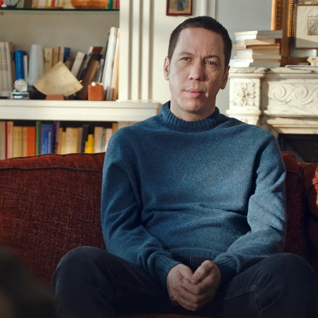 L'image montre un homme assis sur un canapé orange. Il porte un pull bleu et a une expression réfléchie. En arrière-plan, on peut voir une bibliothèque remplie de livres et quelques objets décoratifs. La lumière naturelle entre par une fenêtre, créant une ambiance chaleureuse et confortable dans la pièce. L'homme semble être en train d'écouter ou de participer à une conversation.
