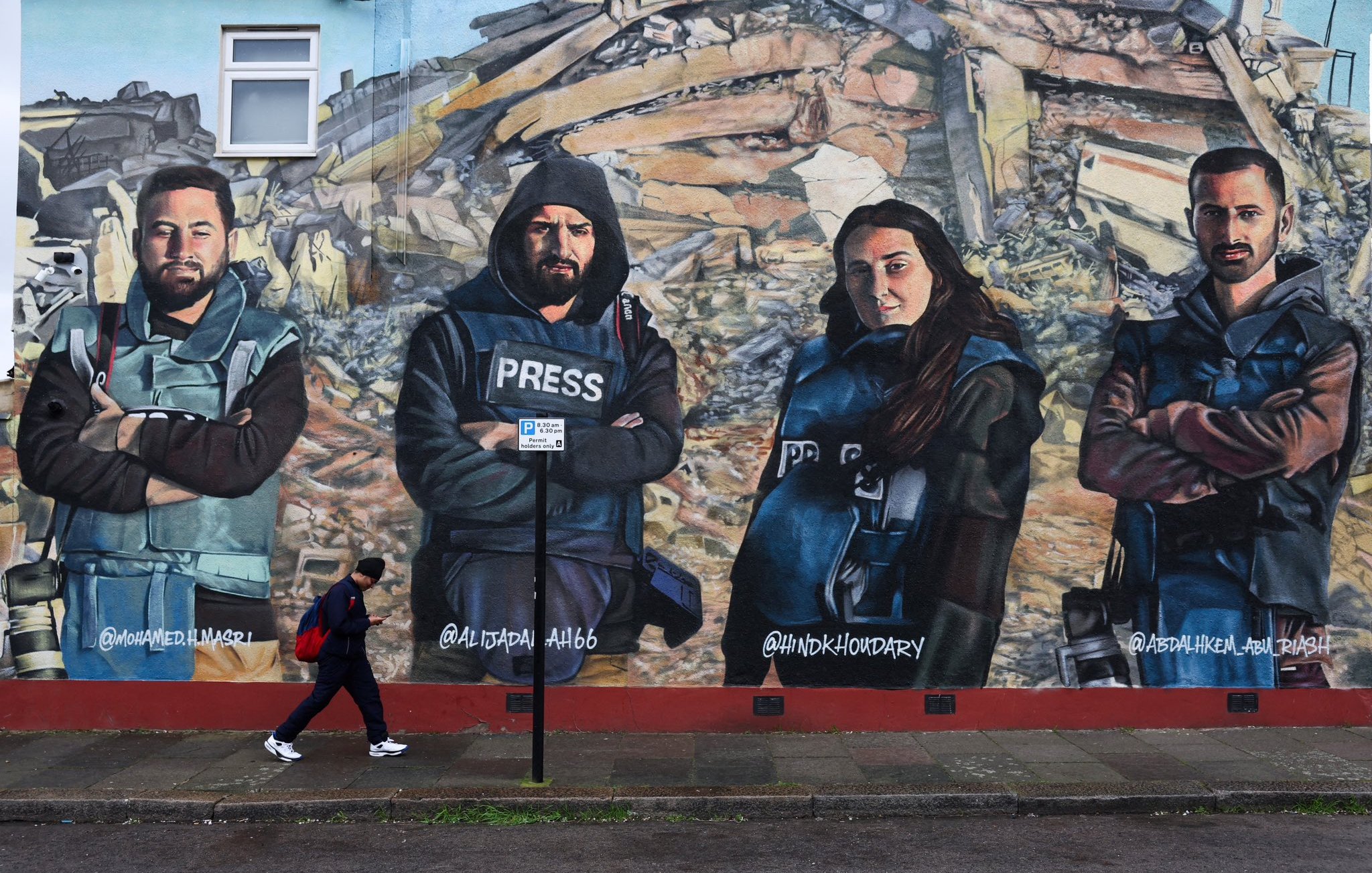 L'immagine mostra un murale che ritrae quattro giornalisti con abbigliamento di protezione e giubbotti con la scritta "PRESS". Sono posizionati in modo prominente davanti a un contesto di distruzione, simile a un'area colpita da un terremoto. Una persona sta camminando lungo il marciapiede di fronte al murale. I volti dei giornalisti esprimono determinazione e serietà, sottolineando il loro lavoro e l'importanza della libertà di stampa.