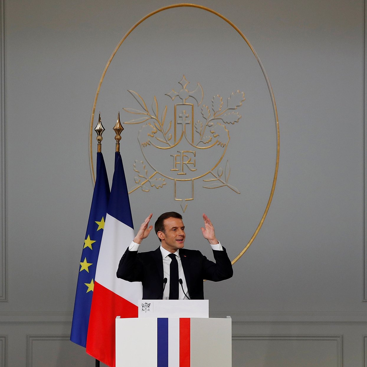 L'image montre un homme debout derrière un pupitre, avec les mains levées comme s'il s'adressait à un auditoire. Derrière lui, il y a un drapeau français et un symbole décoratif sur le mur. Le pupitre présente des couleurs bleu, blanc et rouge, rappelant celles du drapeau français. L'intérieur semble officiel, avec des murs clairs et une ambiance formelle. L'homme semble être en train de faire un discours ou une déclaration importante.