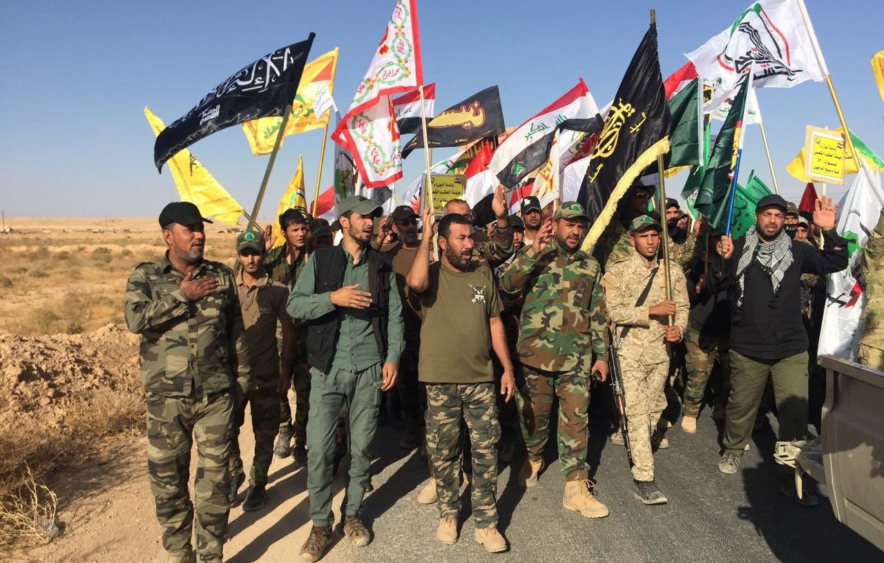 L'image montre un groupe de personnes participant à une manifestation ou une procession. Elles portent des uniformes militaires et tiennent des drapeaux de différentes couleurs et motifs, représentant probablement des groupes ou des mouvements politiques ou religieux. Le paysage environnant est désertique, avec un ciel bleu clair. Les participants semblent engagés et déterminés, marchant ensemble sur une route.