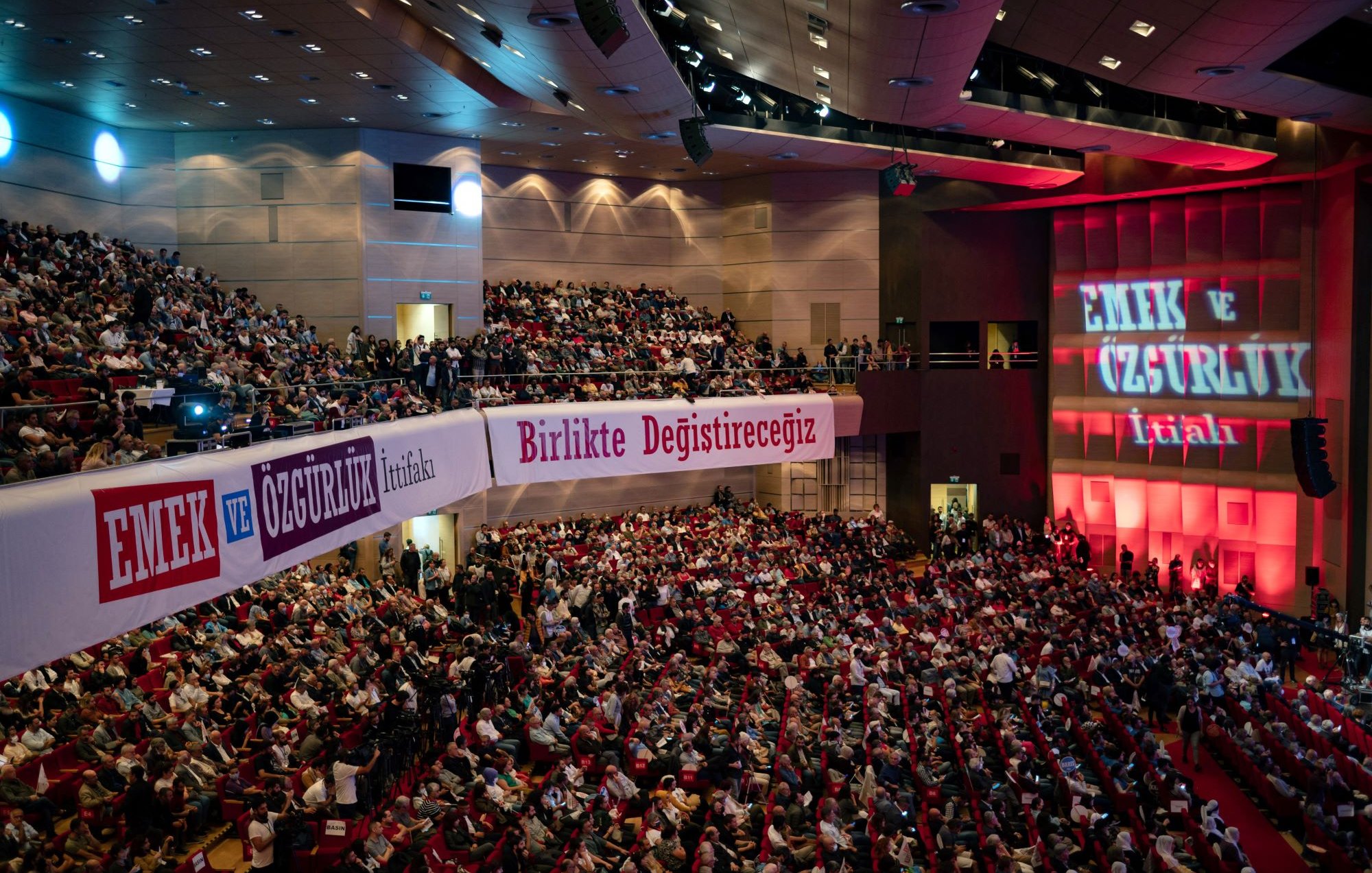 L'image montre une grande salle de conférence ou de réunion, remplie de personnes assises qui semblent assister à un événement. Les spectateurs sont disposés sur des sièges rouges, et des banderoles avec des inscriptions en turc sont accrochées sur les murs, indiquant probablement des slogans liés à des thèmes de travail ou de liberté. Un éclairage rouge illumine le fond de la scène, où des mots supplémentaires sont projetés, renforçant l'atmosphère d'un rassemblement important.