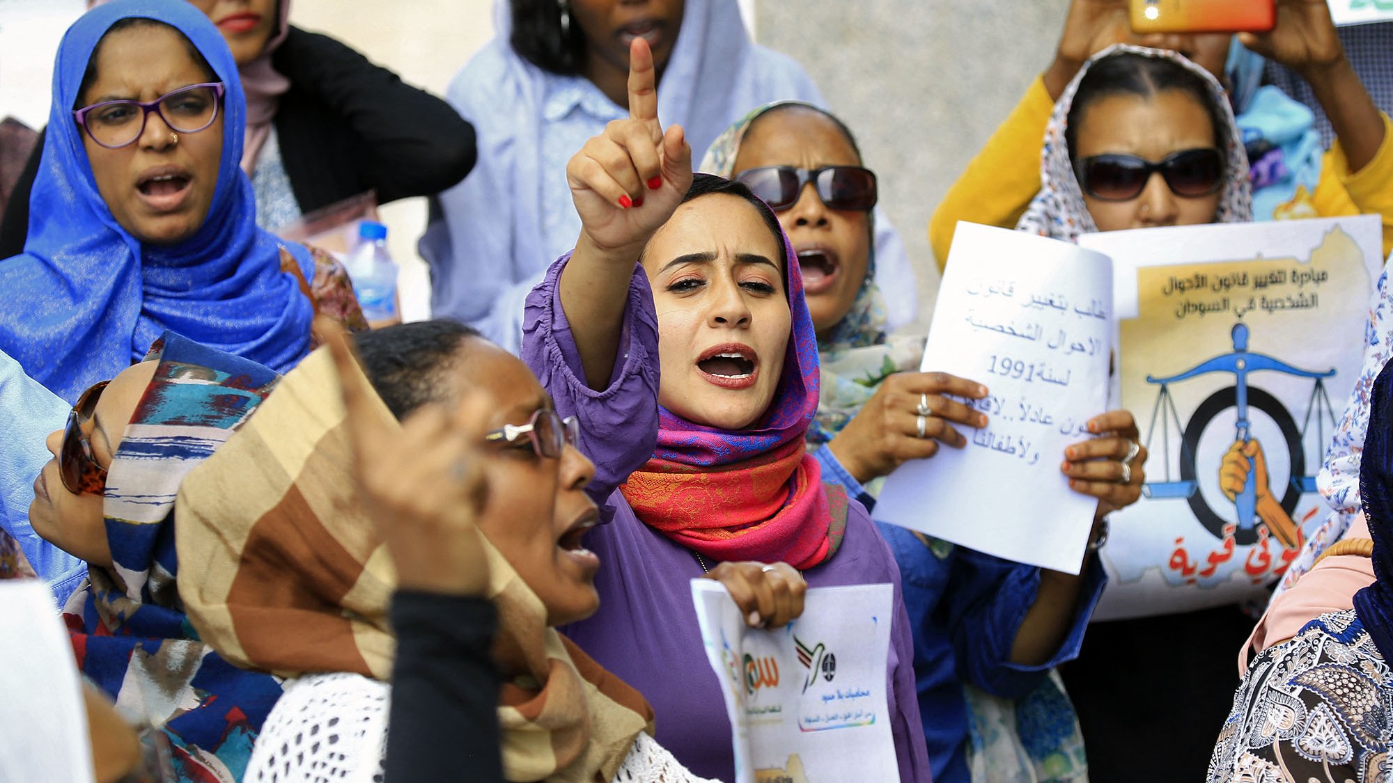 L'image montre un groupe de femmes lors d'une manifestation. Elles portent des foulards et des vêtements colorés. Certaines personnes lèvent les mains et expriment des slogans avec une grande énergie. Des pancartes sont visibles, portant des messages concernant des droits ou des revendications sociales. L'ambiance est dynamique et engagée, avec un fort esprit de solidarité parmi les participantes.