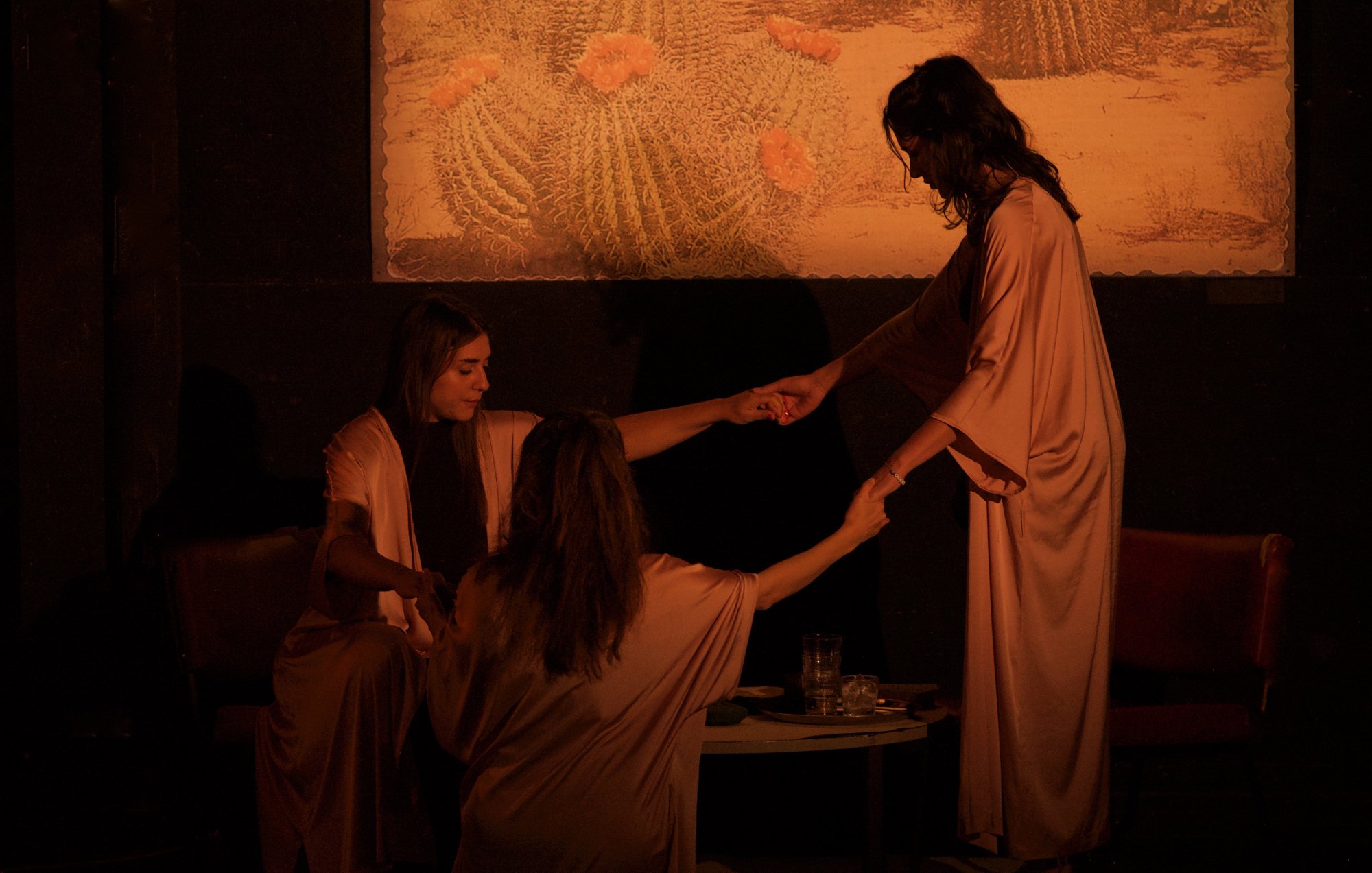 L'image présente une scène théâtrale ou artistique, où trois femmes en vêtements fluides de couleur beige se tiennent autour d'une table basse. Elles semblent engagées dans une interaction attentive, tenant les mains les unes des autres. La lumière est tamisée, créant une atmosphère intime et mystérieuse. En arrière-plan, il y a un tableau représentant des cactus dans un paysage désertique, ajoutant une touche de nature à la scène. Les expressions des femmes ne sont pas clairement visibles, contribuant à l'ambiance intrigante de l'image.