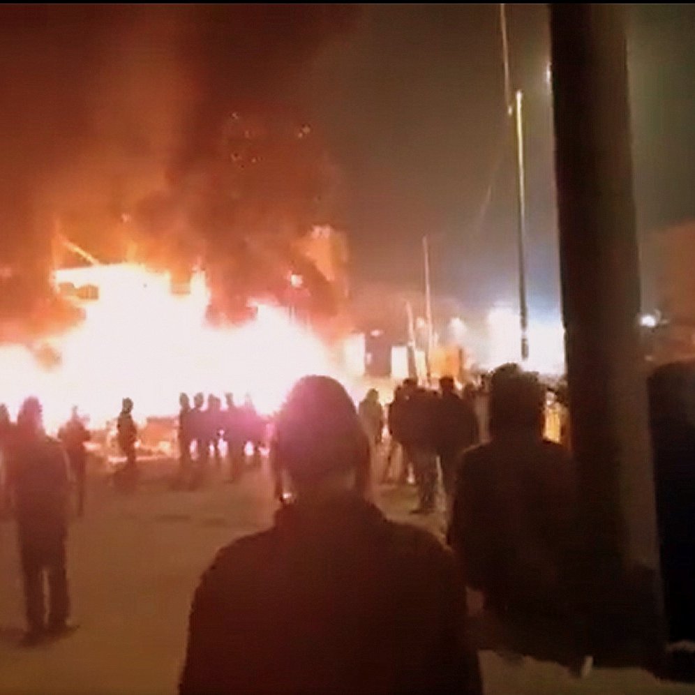 در این تصویر، ظاهراً افرادی در یک صحنه شبانه به سوی یک آتش بزرگ در حال تماشای آن هستند. شعله‌های آتش به شدت در حال سوزاندن یک ساختمان یا ساختار دیگر هستند و دود سیاهی به سمت آسمان بلند می‌شود. مردم در گروه‌های مختلف در پس‌زمینه ایستاده‌اند و به سمت محل آتش نگاه می‌کنند. احتمالاً این صحنه به یک حادثه یا اعتراض مربوط می‌شود. روشنایی آتش فضای اطراف را درخشان کرده است.