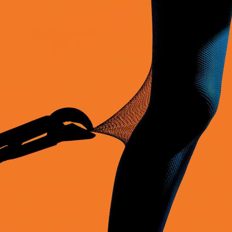 L'image montre une silhouette humaine, probablement une jambe, portant des collants en filet. Un outil (possiblement une pince) semble tirer sur le collant, créant une tension visuelle intéressante. En arrière-plan, une couleur orange vive attire l'œil et crée un contraste frappant avec la silhouette sombre. L'ensemble évoque une sensation de dynamisme et d'innovation.