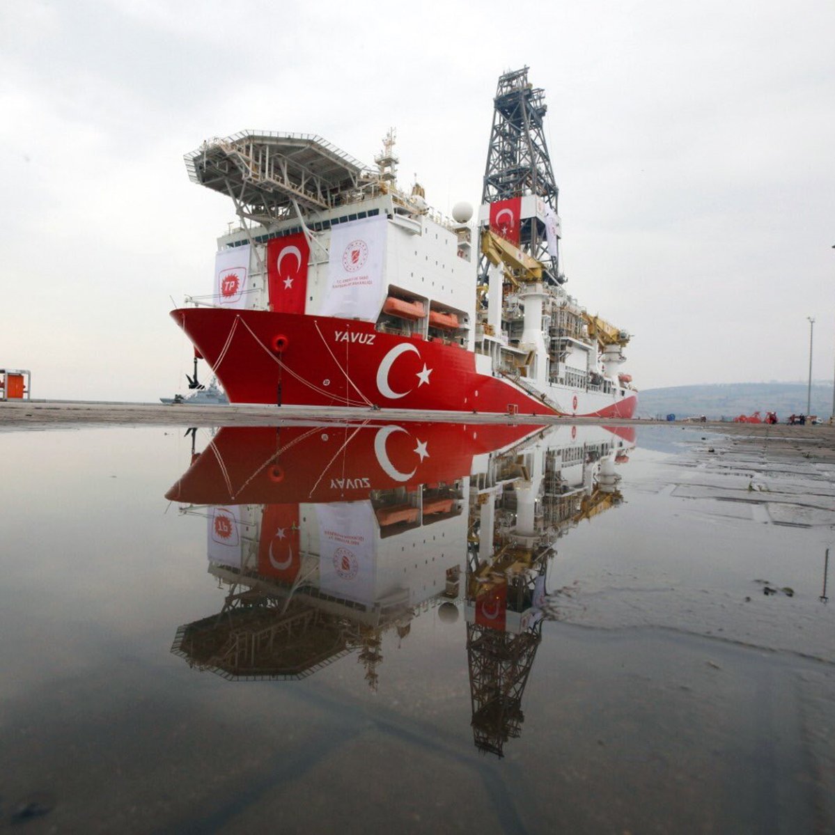 L'image montre un navire de forage maritime, nommé "Yavuz", qui est amarré au port. Le bateau arbore des couleurs dominantes de rouge et blanc, représentant le drapeau turc. On peut également voir des équipements de forage sur le pont du navire. Dans la partie inférieure de l'image, il y a des réflexions du navire dans une flaque d'eau sur le sol. Le ciel en arrière-plan est nuageux, créant une atmosphère calme.