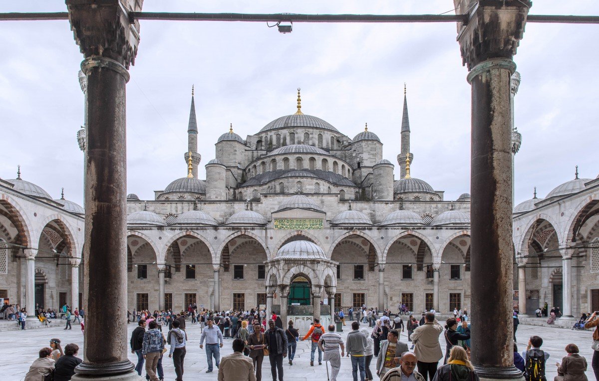 The image features a grand architectural view of the Sultan Ahmed Mosque, commonly known as the Blue Mosque, in Istanbul, Turkey. In the foreground, you can see a bustling courtyard filled with visitors and tourists. The mosque's impressive structure is visible in the background, characterized by its large dome and multiple tall minarets reaching upward. The scene is framed by arched columns, adding to the architectural beauty. The sky appears overcast, creating a soft light that enhances the details of the mosque's intricate design.