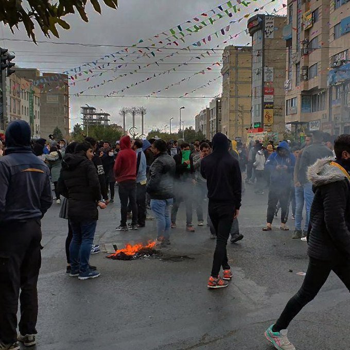 تظهر الصورة مجموعة كبيرة من الناس في منطقة حضرية، حيث يبدو أن هناك تظاهرة أو تجمع حاشد. يتواجد المتظاهرون في الشارع، وبعضهم يرتدي أقنعة وجه. هناك دخان يتصاعد من نيران صغيرة مشتعلة على الأرض. السماء تبدو غائمة، وهناك زينة ملونة معلقة في الجو، مما يشير إلى وجود نشاطات أو احتفالات بجانب المظاهرات. الأجواء تعكس التوتر نتيجة الظاهر.