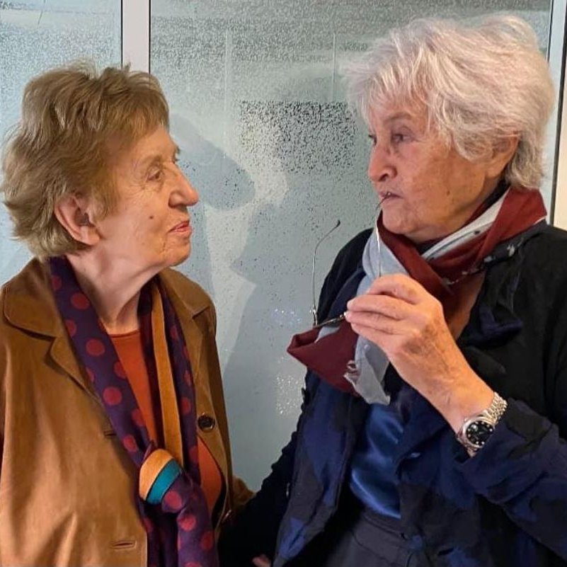 L'image montre deux femmes âgées engagées dans une conversation. Elles semblent amicales et souriantes. L'une porte une veste marron avec un foulard à pois, tandis que l'autre est habillée de manière similaire, avec une écharpe. L'arrière-plan est flou, mettant en valeur leur interaction.