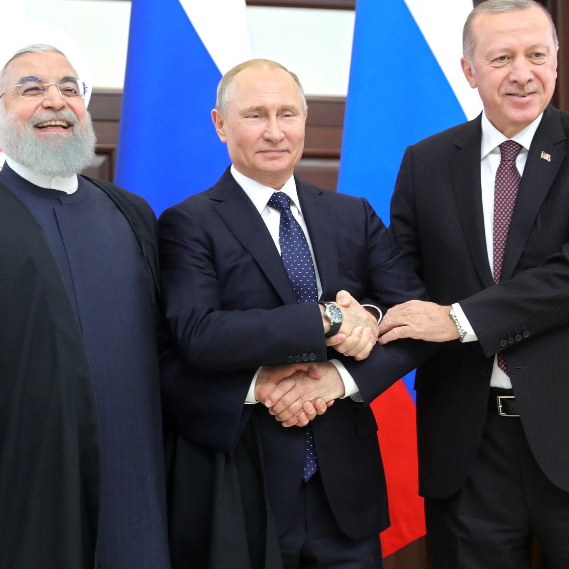 L'image montre trois dirigeants se tenant côte à côte. À gauche, un homme portant une robe sombre et une barbe grise, au centre un homme en costume sombre avec une cravate, et à droite, un homme également en costume qui semble sourire. Ils se tiennent par les bras, affichant une expression amicale, devant des drapeaux représentant leurs pays respectifs. L'environnement semble être une salle de réunion ou une conférence.