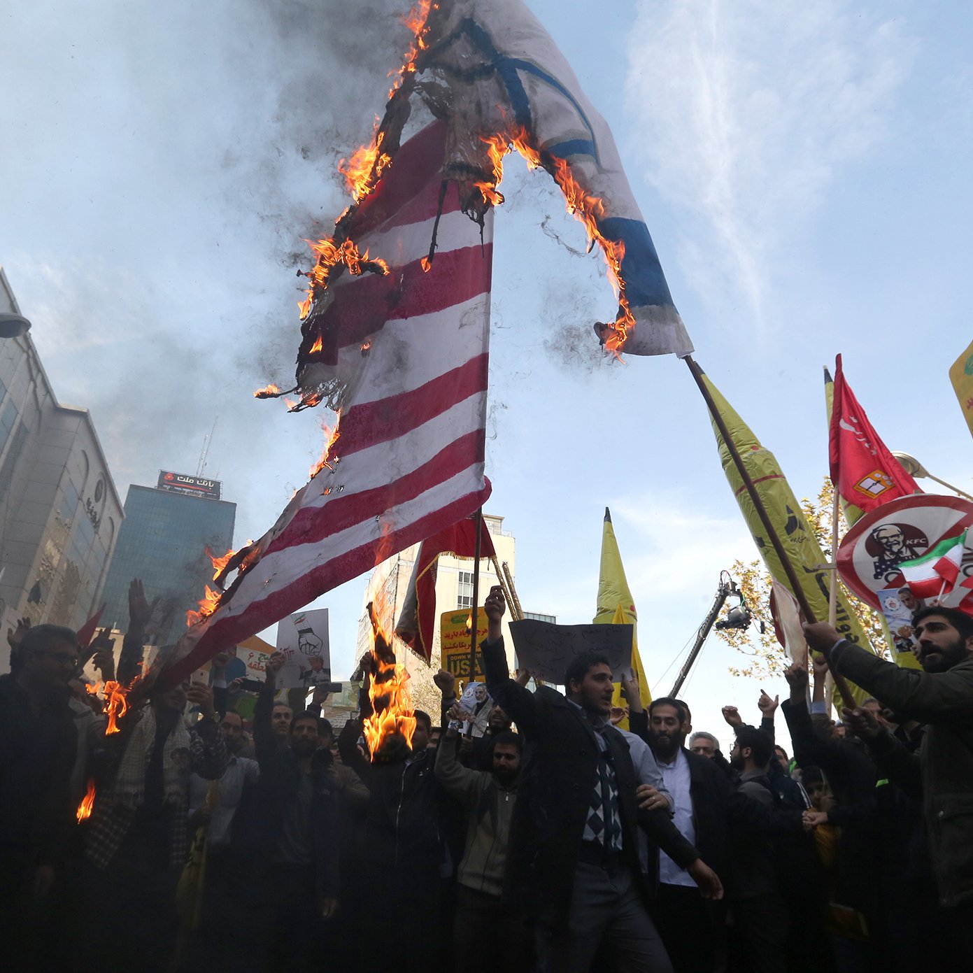 L'image montre une scène de manifestation où des personnes sont rassemblées pour protester. Au centre, un drapeau semble être en feu, ce qui attire l'attention des manifestants. Ils tiennent également des pancartes avec différents slogans et symboles. L'atmosphère est tendue, et on ressent un fort sentiment d'engagement parmi les participants. L'arrière-plan présente des bâtiments urbains, suggérant une grande ville.