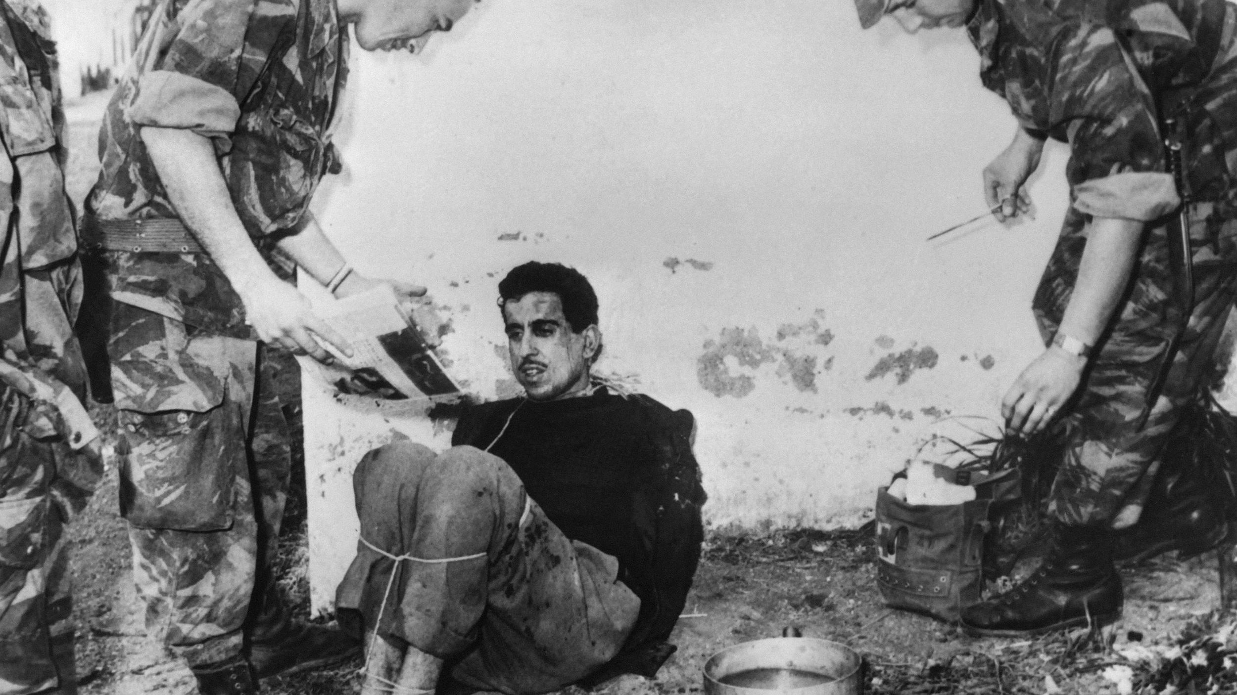 L'image montre un homme assis et attaché, visiblement en détresse, tandis que deux soldats, habillés en uniforme militaire, se tiennent près de lui. Un des soldats semble examiner un document ou une photographie, tandis que l'autre s'approche avec un équipement. L'environnement est sobre, et l'ambiance générale de la scène dégage un sentiment de tension.