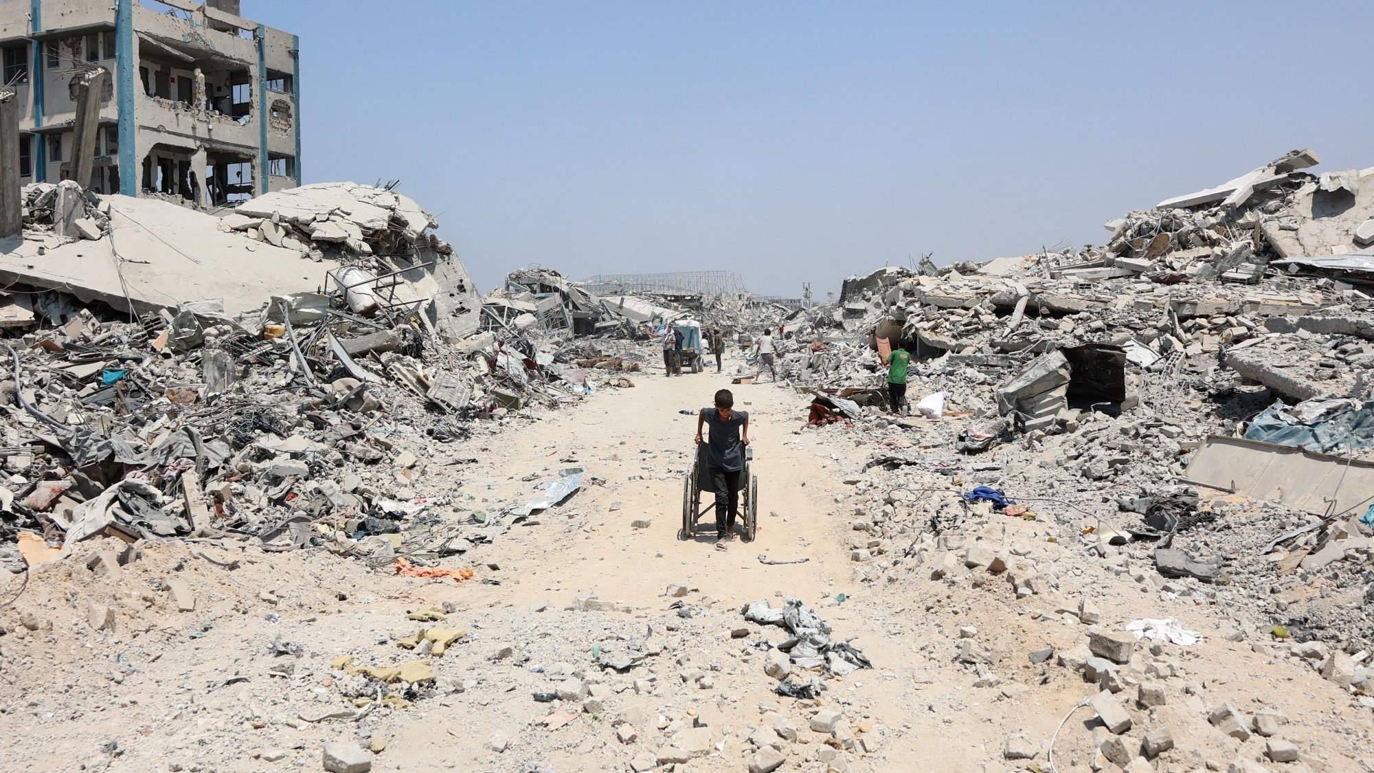 Una strada polverosa tra macerie di edifici distrutti, con una persona in sedia a rotelle.