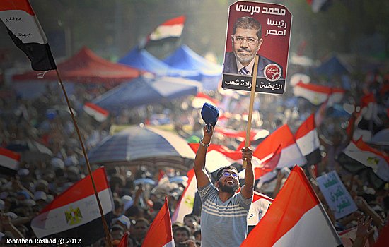 L'image montre une grande foule de manifestants brandissant des drapeaux égyptiens. Au premier plan, un homme lève une pancarte avec le portrait de Mohamed Morsi, qui était le président de l'Égypte. Les participants semblent enthousiasmés et mobilisés, reflétant une atmosphère de rassemblement populaire et d'engagement politique. Le paysage est rempli de tentes et d'une multitude de personnes, créant une impression de solidarité.