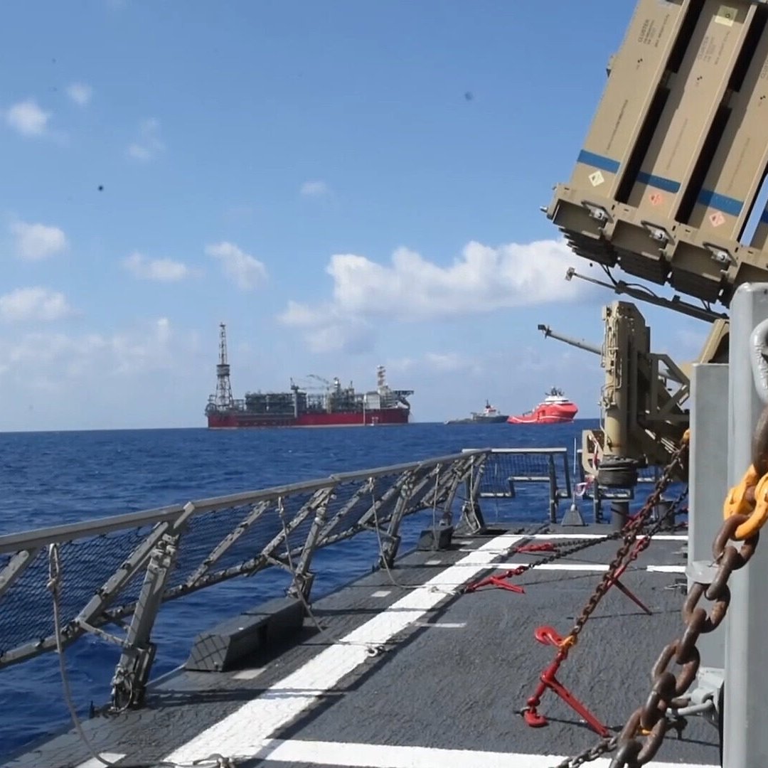 L'image montre un navire en mer, avec des structures de forage pétrolier visibles à l'horizon. On peut apercevoir un système de défense intégrée, probablement dédié à la surveillance ou à la protection, installé sur le pont du navire. Le ciel est clair avec quelques nuages, et la mer est calme, créant une atmosphère maritime paisible. Des chaînes et des équipements de sécurité sont également visibles sur le pont.