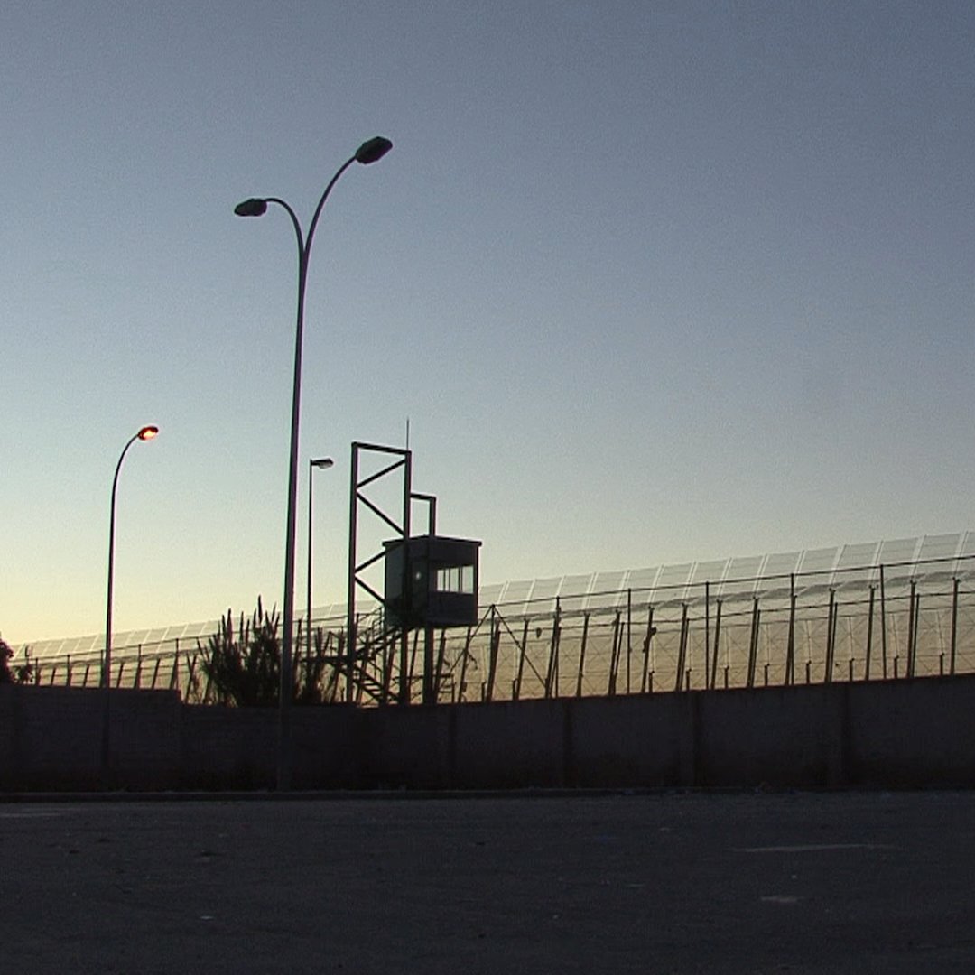 L'image montre une clôture de sécurité, probablement d'une prison ou d'une zone contrôlée. On aperçoit une tour d'observation au fond, illuminée par des lampadaires. La scène se déroule au crépuscule, avec un ciel qui passe des teintes orange et bleues, créant une atmosphère à la fois calme et tendue. Le mur en béton à la base de la clôture souligne le caractère restrictif de l'endroit.