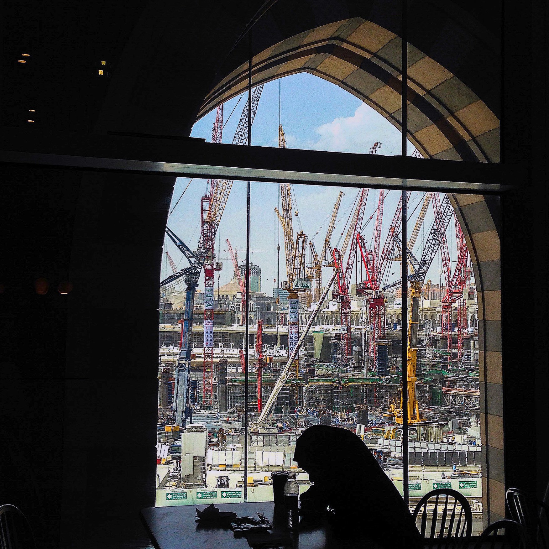 L'image montre un chantier de construction vu à travers une grande fenêtre à arc. À l'intérieur, on aperçoit une silhouette assise à une table. De l'autre côté de la vitre, de nombreux grues et engins de chantier se dressent, travaillant à un projet de grande envergure. Le ciel est partiellement dégagé, ajoutant une touche de lumière à la scène. L'ensemble dégage une atmosphère de travail intensif et de développement urbain.
