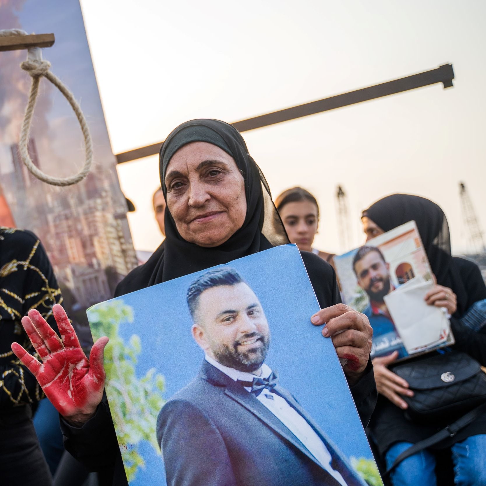 L'image montre un rassemblement commémoratif. Au premier plan, deux femmes sont présentes. L'une porte un hijab noir et tient une grande photo d'un homme. L'autre femme, à sa gauche, porte un masque bleu et semble également participer à la manifestation. La femme avec la photo a une main teintée de rouge, symbolisant peut-être la douleur ou la protestation. En arrière-plan, on peut voir des personnes rassemblées, ainsi qu'un décor urbain qui suggère un contexte de tension ou de lutte.