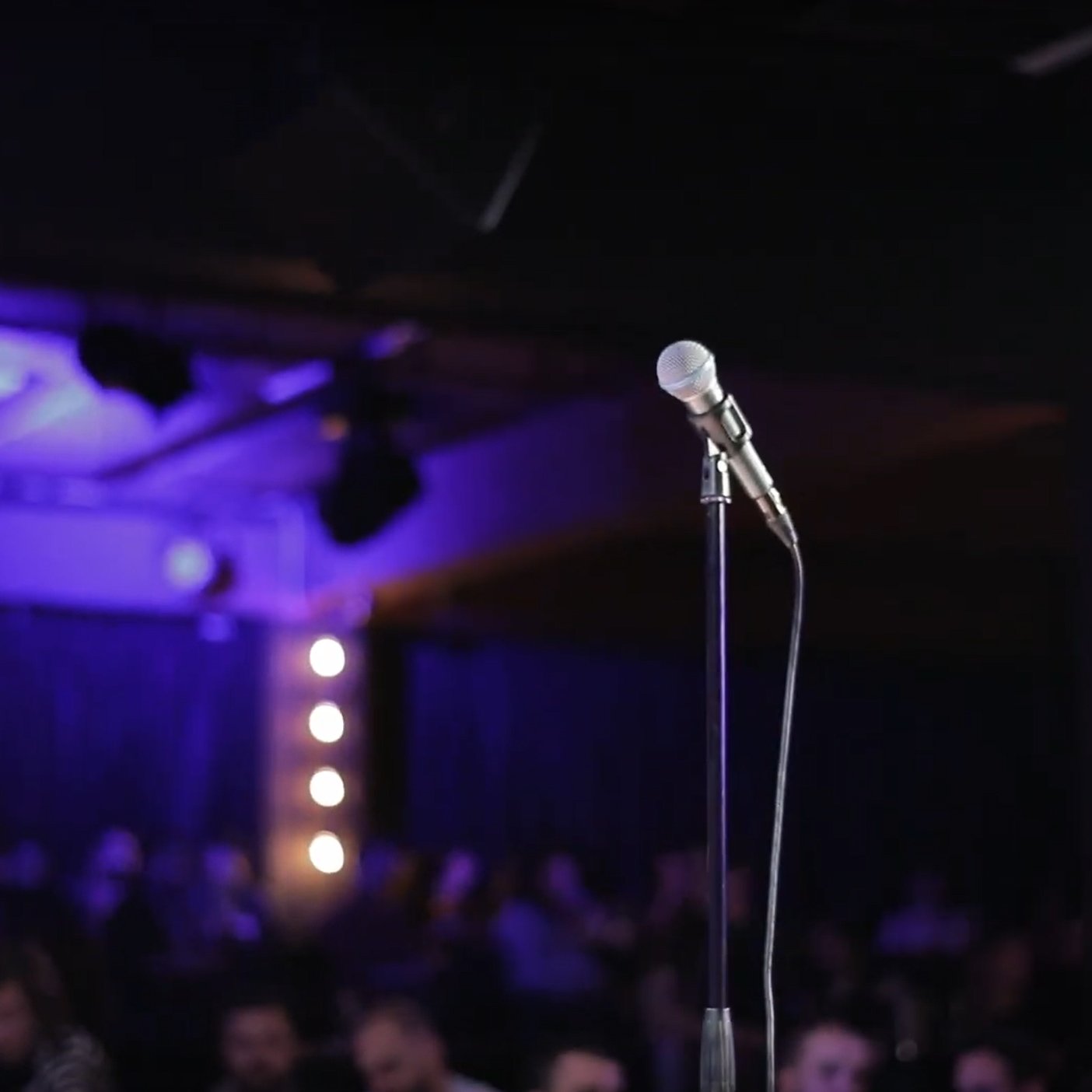 L'image montre un microphone sur un pied, au premier plan, sur une scène sombre. À l'arrière-plan, on aperçoit une audience floue et des lumières tamisées qui créent une ambiance de spectacle.