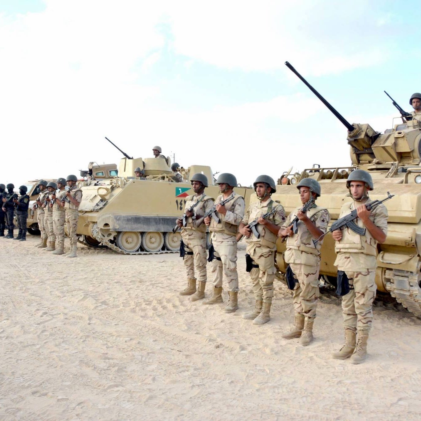 L'image montre une formation militaire dans un environnement désertique. Au premier plan, des soldats en uniforme, armés et en position de garde, se tiennent alignés. En arrière-plan, on aperçoit plusieurs véhicules blindés, dont certains sont équipés de tourelles. Le ciel est clair, et le sol est sablonneux, typique des zones désertiques. Les soldats semblent concentrés et prêts pour une mission.