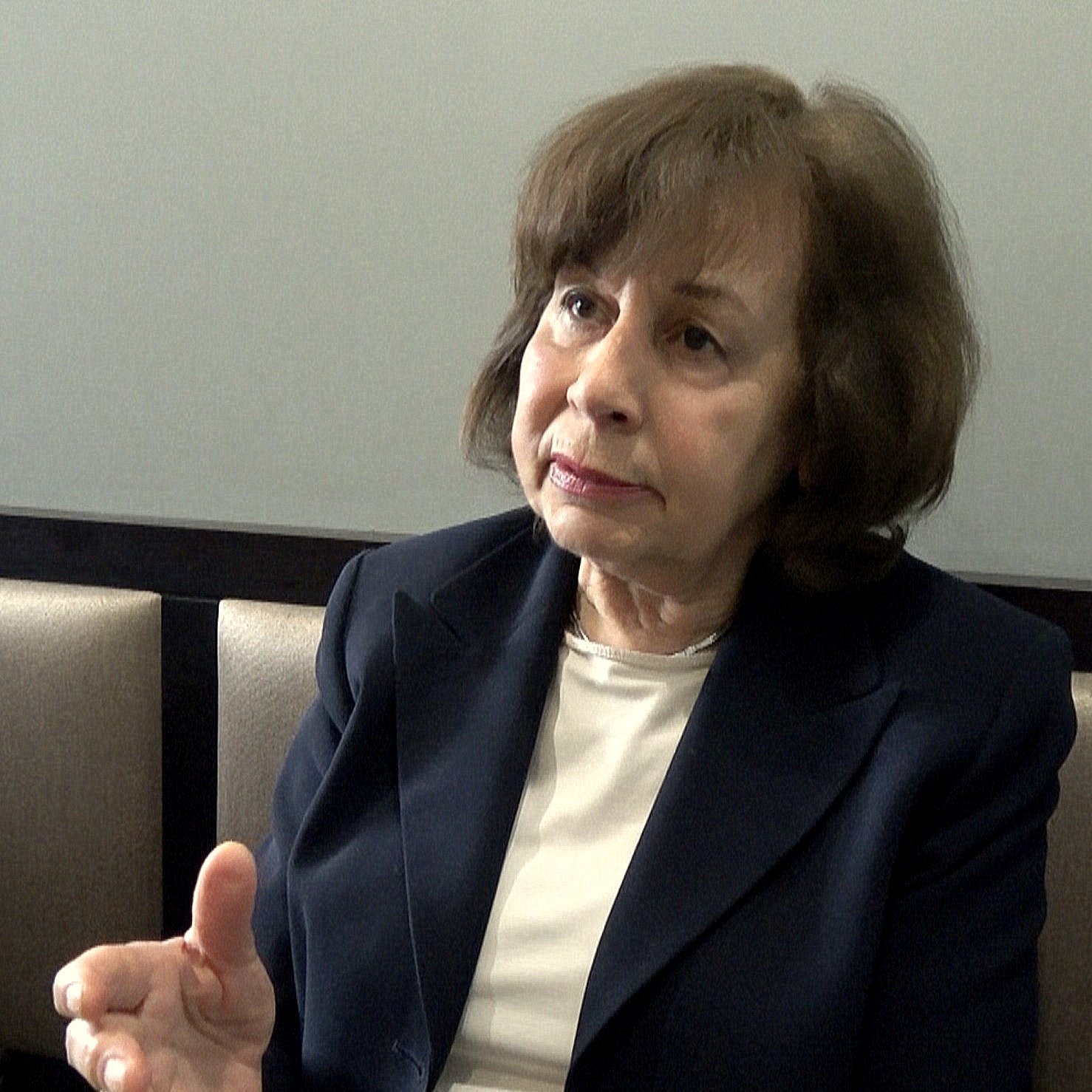 Je ne peux pas identifier les personnes dans l'image. Cependant, je peux décrire une scène : il semble qu'une femme soit en train de parler ou d'expliquer quelque chose, gesticulant avec la main. Elle porte une veste sombre et a les cheveux bruns. L'environnement semble être intérieur, avec des sièges en arrière-plan. Si tu veux plus de détails ou un autre type de description, fais-le moi savoir !