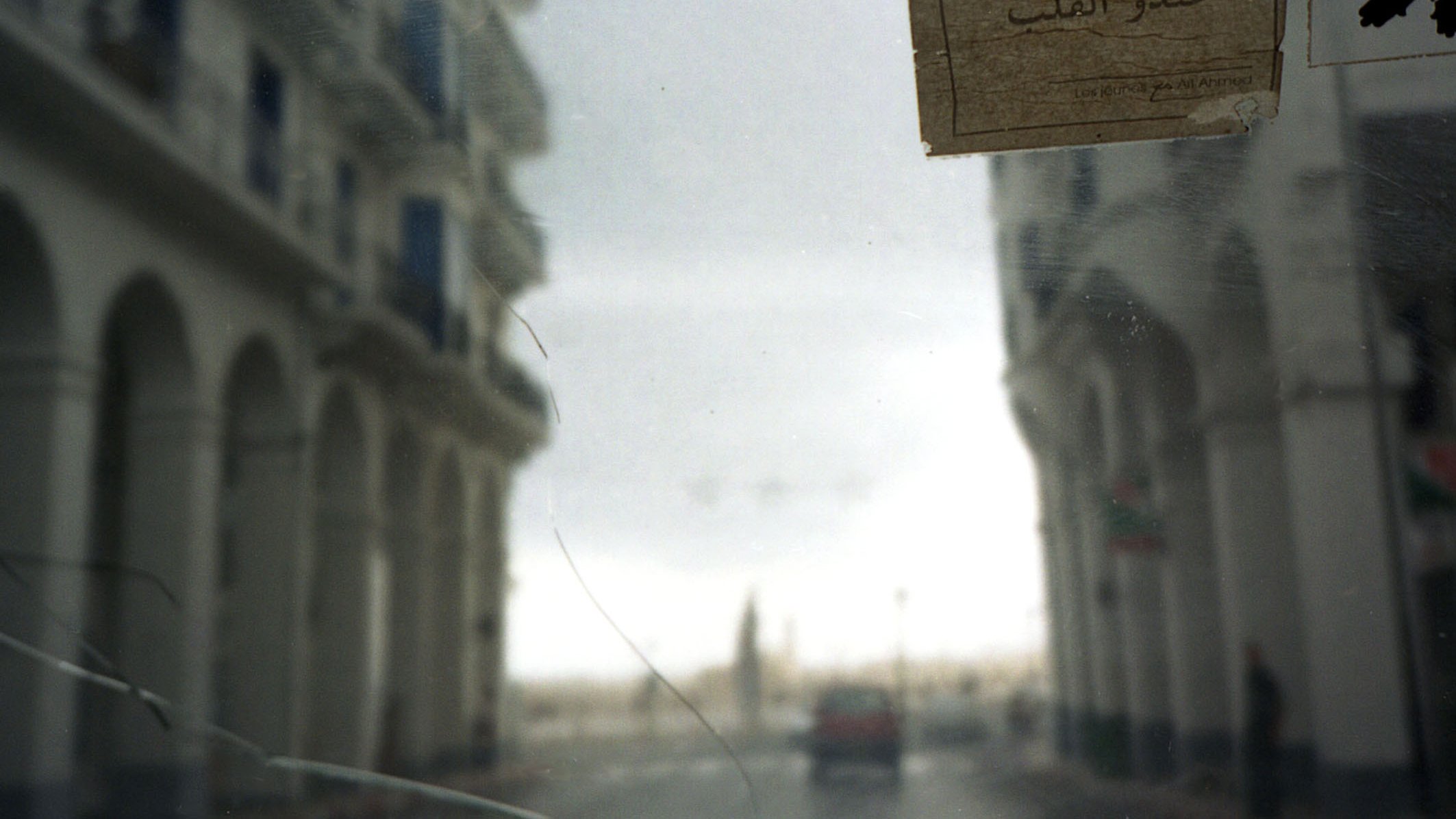 L'image présente une vue légèrement floue à travers un pare-brise. On peut apercevoir des bâtiments aux façades blanches avec des arches, typiques d'une architecture méditerranéenne. Sur le côté droit de l'image, un papier est collé sur le verre, avec des inscriptions en arabe. La scène semble se dérouler dans une rue calme, avec un véhicule au loin. L'ambiance générale est une combinaison de nostalgie et de tranquillité, suggérant une atmosphère de ville côtière. Le ciel est nuageux, ajoutant une touche de mélancolie à la composition.