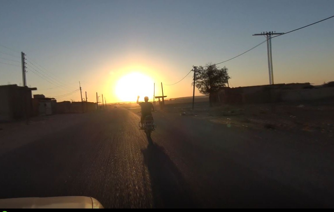 L'image montre un paysage rural au crépuscule. On voit une route désertique s'étendant devant, avec un motard roulant vers la direction du soleil couchant. Les rayons du soleil illuminent le ciel, créant une ambiance chaleureuse. De chaque côté de la route, on aperçoit quelques bâtiments et des poteaux électriques. L'atmosphère semble paisible et tranquille.