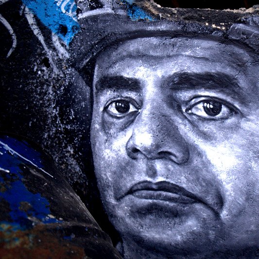 L'image représente une œuvre de street art, probablement une peinture murale, qui montre un portrait en noir et blanc d'un homme. Le visage est détaillé, avec une expression sérieuse et des yeux perçants. Le fond semble être un mur abîmé, avec des graffitis et des marques de peinture colorées, ce qui crée un contraste avec le portrait monochrome. L'ensemble donne une impression de profondeur et de caractère à l'œuvre.
