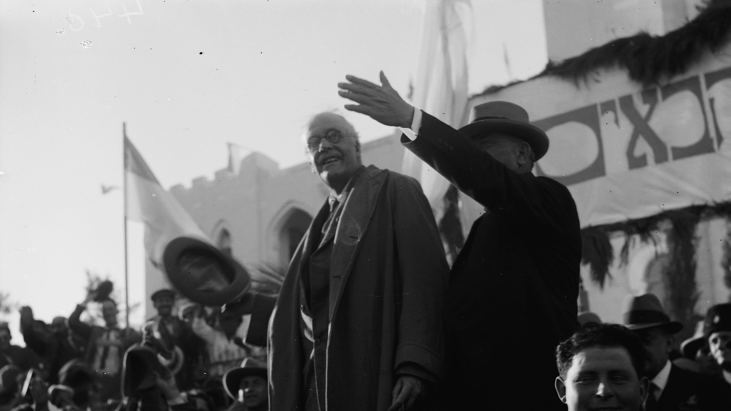 در این تصویر، دو مرد در حال سخنرانی یا معرفی هم دیگر هستند. یکی از آن‌ها به وضوح در حال صحبت کردن است و دستش را به علامت اشاره به سمت جمعیت بالا برده است. مرد دیگر کنار او ایستاده و به نظر می‌رسد که به جمعیت توجه دارد. پس‌زمینه تصویر ازدحام جمعیت را نشان می‌دهد که در حال تماشای آنها هستند. بعضی پرچم‌ها و طراحی‌هایی در پس‌زمینه دیده می‌شوند که احتمالاً به یک رویداد خاص اشاره دارند. این تصویر حال و هوای تاریخی و اجتماعی را به نمایش می‌گذارد.