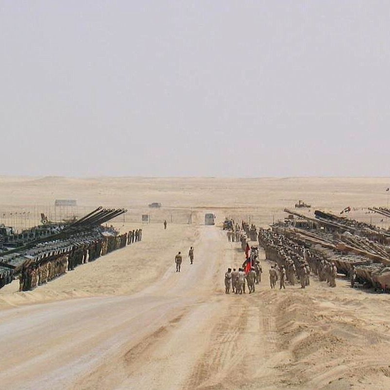 تظهر في الصورة مشهدًا صحراويًا يضم العديد من الدبابات والمركبات العسكرية. يقف عدد من الجنود في المنتصف، بينما تتواجد مركبات أخرى مصفوفة على جانبي الطريق. الخلفية تتكون من رمال الصحراء وأفق بعيد، مما يبرز الطبيعة القاحلة للمكان. تخلق الصورة انطباعًا بالقوة العسكرية والتنظيم.