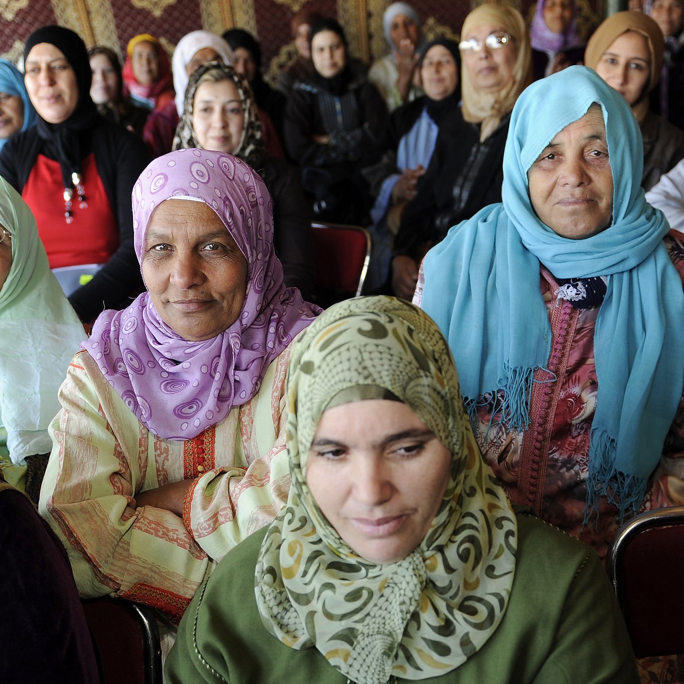 L'image montre un groupe de femmes assises, majoritairement vêtues de vêtements traditionnels et portant des foulards. Certaines portent des vêtements clairs tandis que d'autres ont des tenues plus sombres. Les expressions des femmes varient, allant de souriantes à attentives. L'environnement semble être une réunion ou un rassemblement, avec des murs ornés de motifs. L'atmosphère semble conviviale et solennelle.