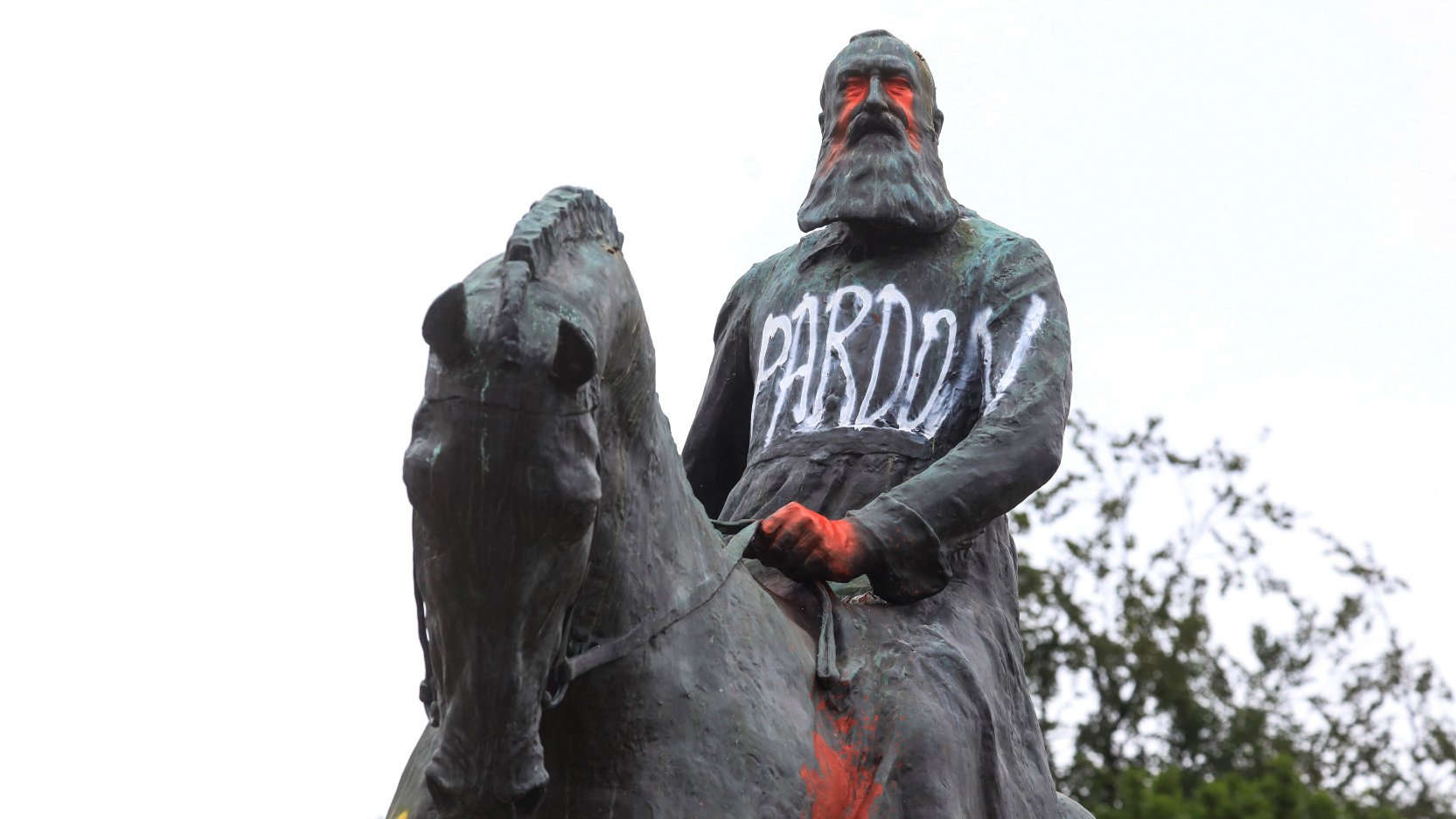 این تصویر نشان‌دهنده یک مجسمه است که یک شخصیت سوار بر اسب را به تصویر کشیده است. مجسمه به نظر می‌رسد که به طور هنری با رنگ‌های مختلف نقاشی شده و روی آن عبارت‌هایی نوشته شده است. کلمات "PARDON" و "RACIST" به وضوح روی مجسمه دیده می‌شوند، که بیانگر اعتراضات یا نظرات انتقادی نسبت به این شخصیت تاریخی است. زمینه تصویر شامل درختان و آسمان ابری است.